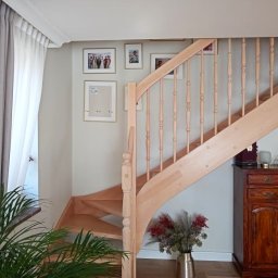 PIO-MAR Glazurnictwo - Jasne drewniane schody z balustradą w domu, pod schodami ciemna drewniana komoda, na ścianie galeria obrazków w złotych ramkach, obok okna z zasłonami i doniczkowa roślina.