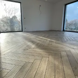 Ułożona w jodełkę podłoga z płytek imitujących drewno w jasnym, pustym pokoju z dużymi oknami i minimalistycznym oświetleniem.