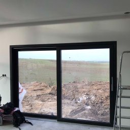 PIO-MAR Glazurnictwo - Wnętrze w trakcie wykańczania: duże przesuwne okno z czarną ramą, widok na pole i ziemię, ściany w surowym stanie, drabina aluminiowa, worki z zaprawą budowlaną i lampy budowlane.