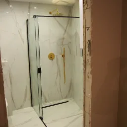 Nowoczesna kabina prysznicowa z czarnymi ramami i złotą armaturą na tle marmurowych płytek, widoczny odpływ liniowy i fragment surowej ściany w tle.