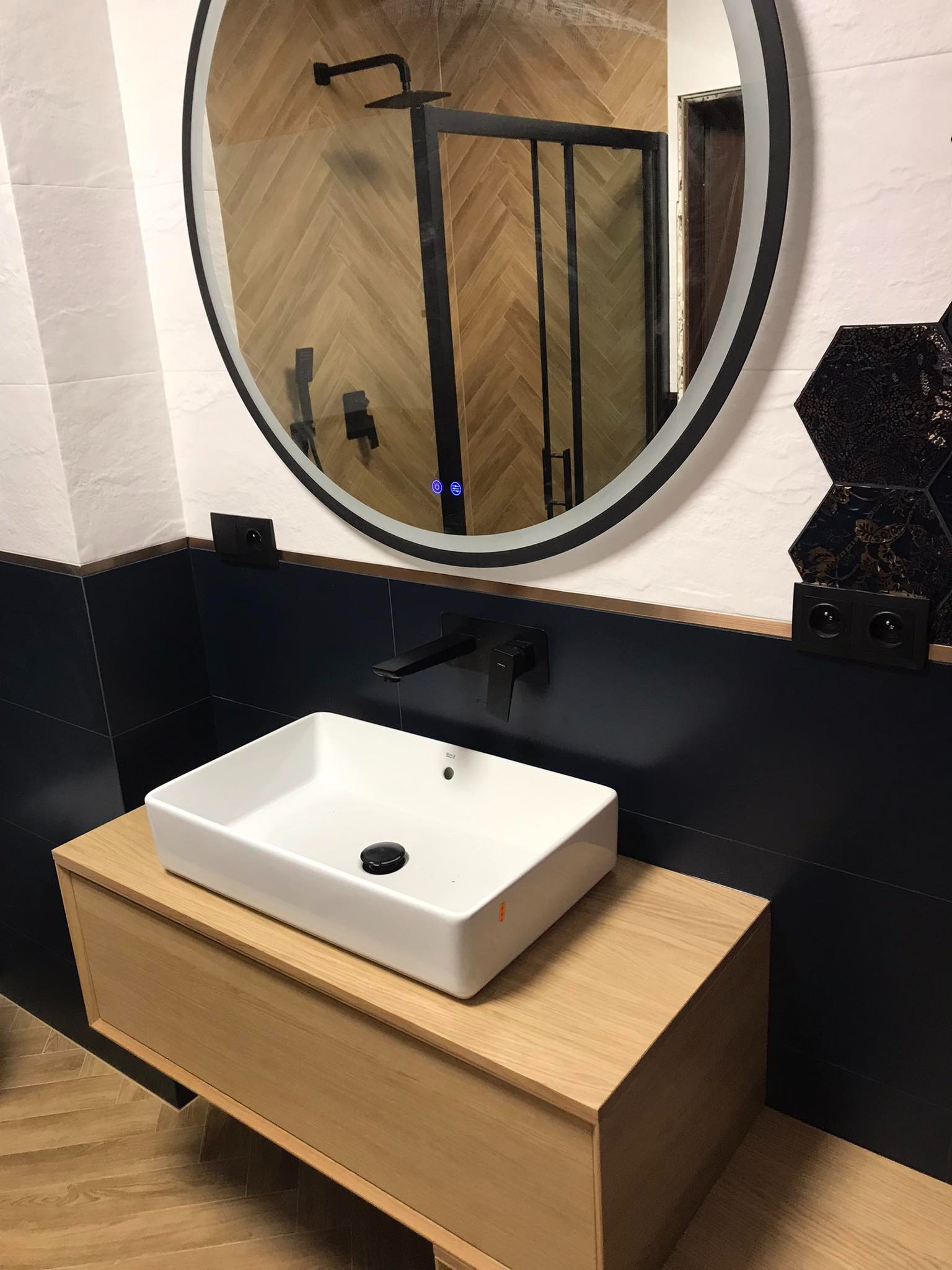 Nowoczesna łazienka z umywalką nablatową, czarną armaturą i okrągłym lustrem z podświetleniem LED, w tle widoczna kabina prysznicowa i płytki ułożone w jodełkę na ścianie.