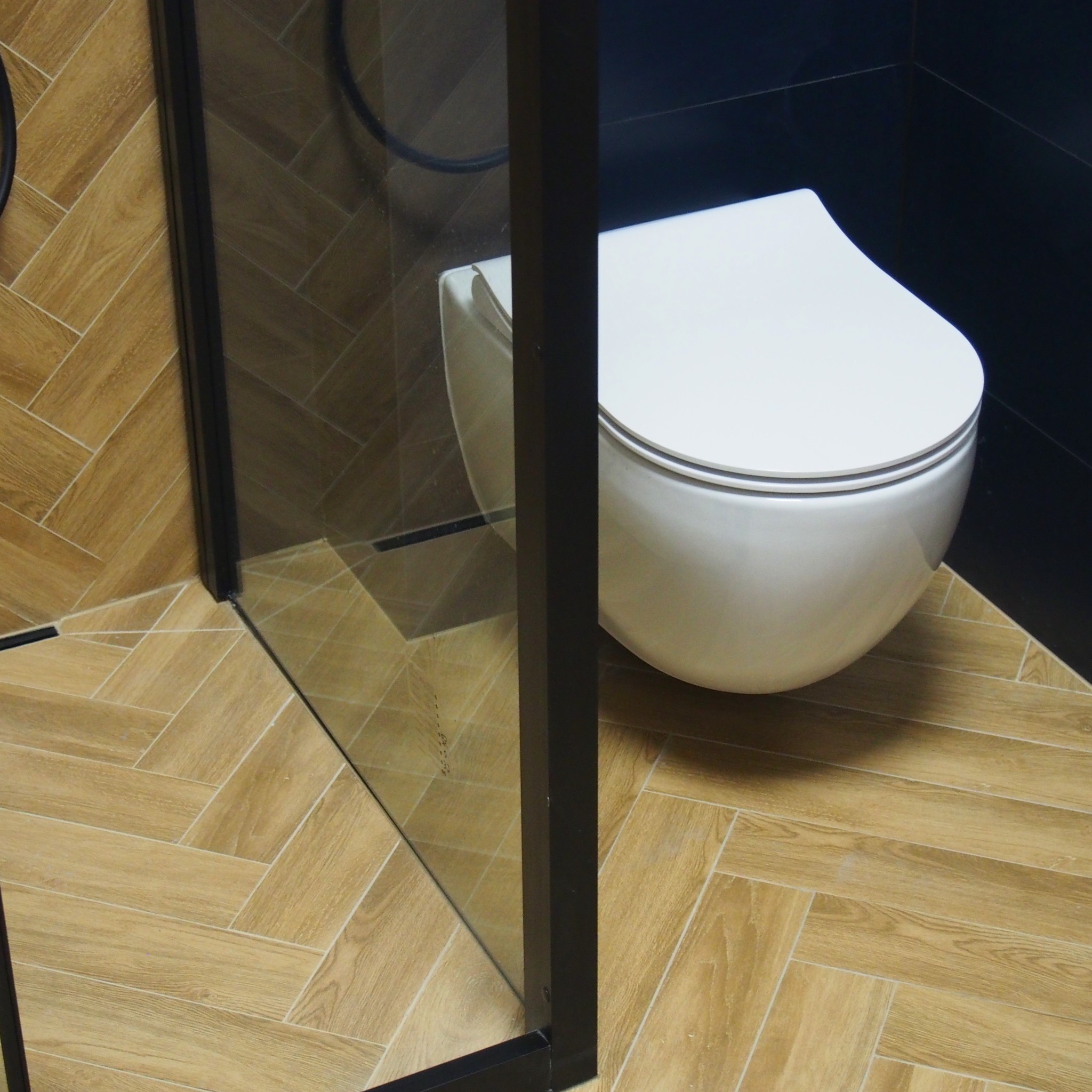 Nowoczesna, podwieszana toaleta w łazience z czarnymi płytkami i podłogą z płytek imitujących drewniany parkiet ułożony w jodełkę, widok przez szklaną ściankę kabiny prysznicowej z czarnymi profilami.