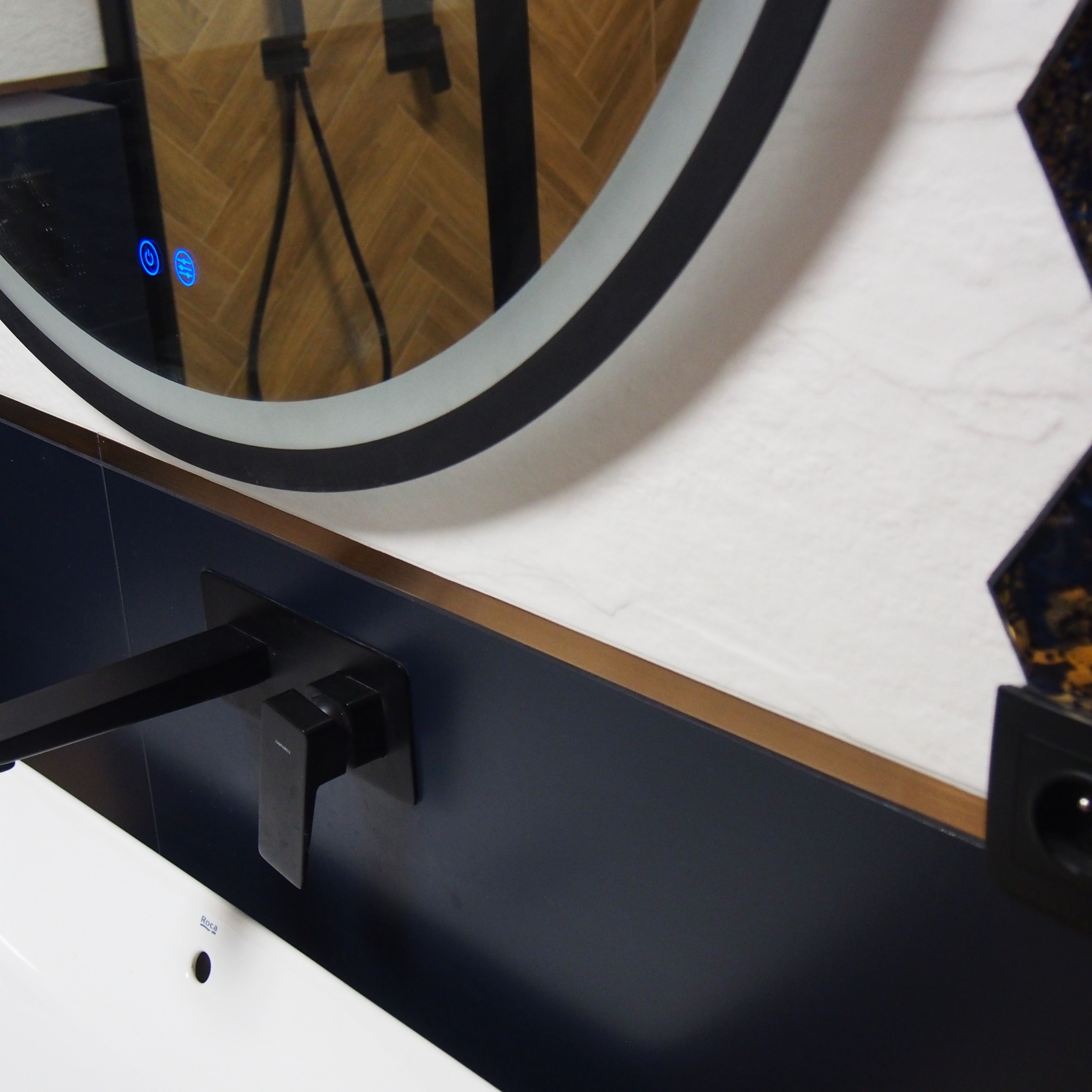 Fragment łazienki z okrągłym lustrem z podświetleniem LED, czarną armaturą i umywalką Roca na tle ściany wykończonej białymi płytkami i elementami dekoracyjnymi.