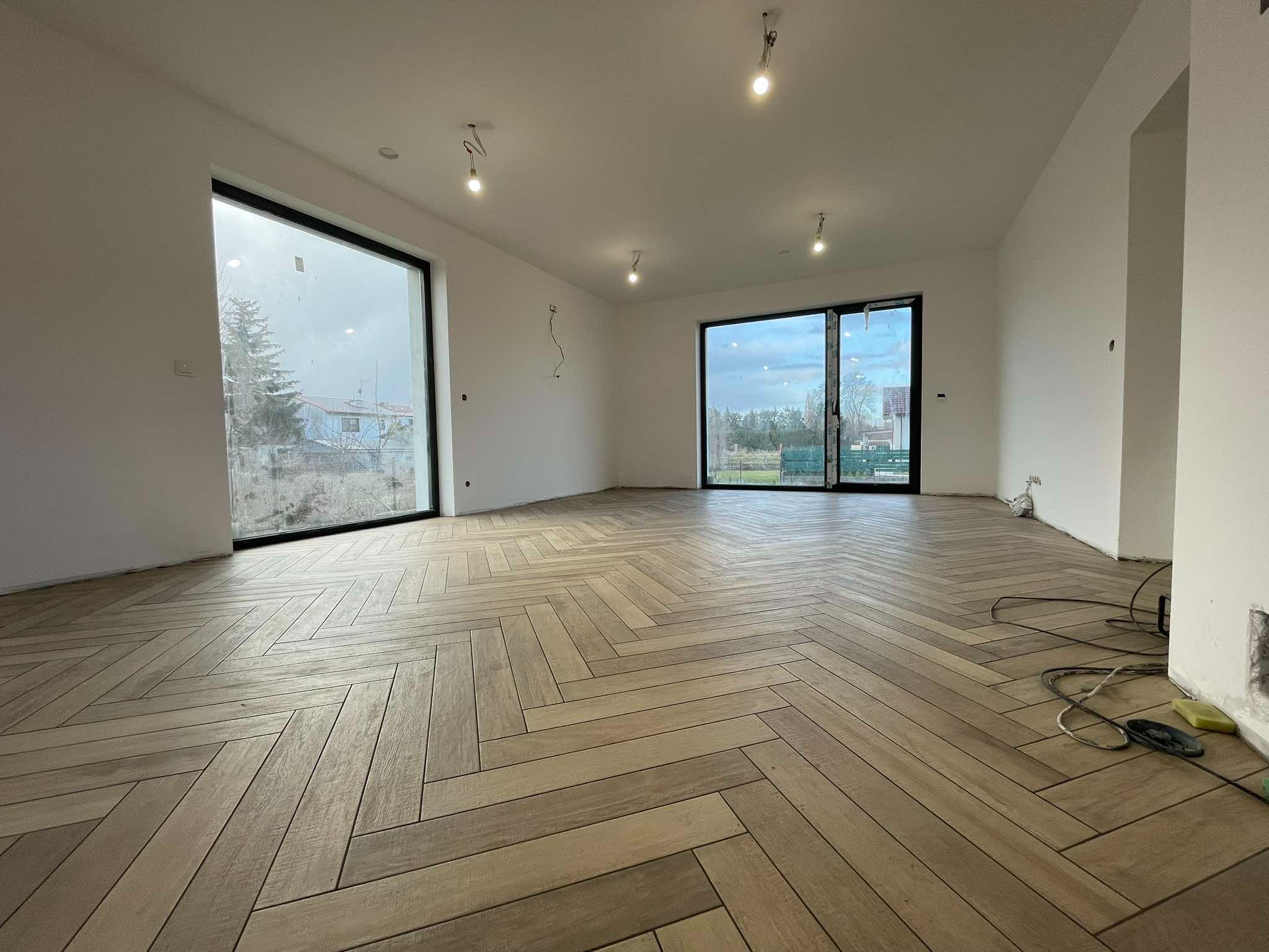 Jasna, przestronna sala z ułożoną w jodełkę podłogą z płytek imitujących drewno, duże okna z czarnymi ramami, surowe ściany i wiszące kable sugerujące prace wykończeniowe.