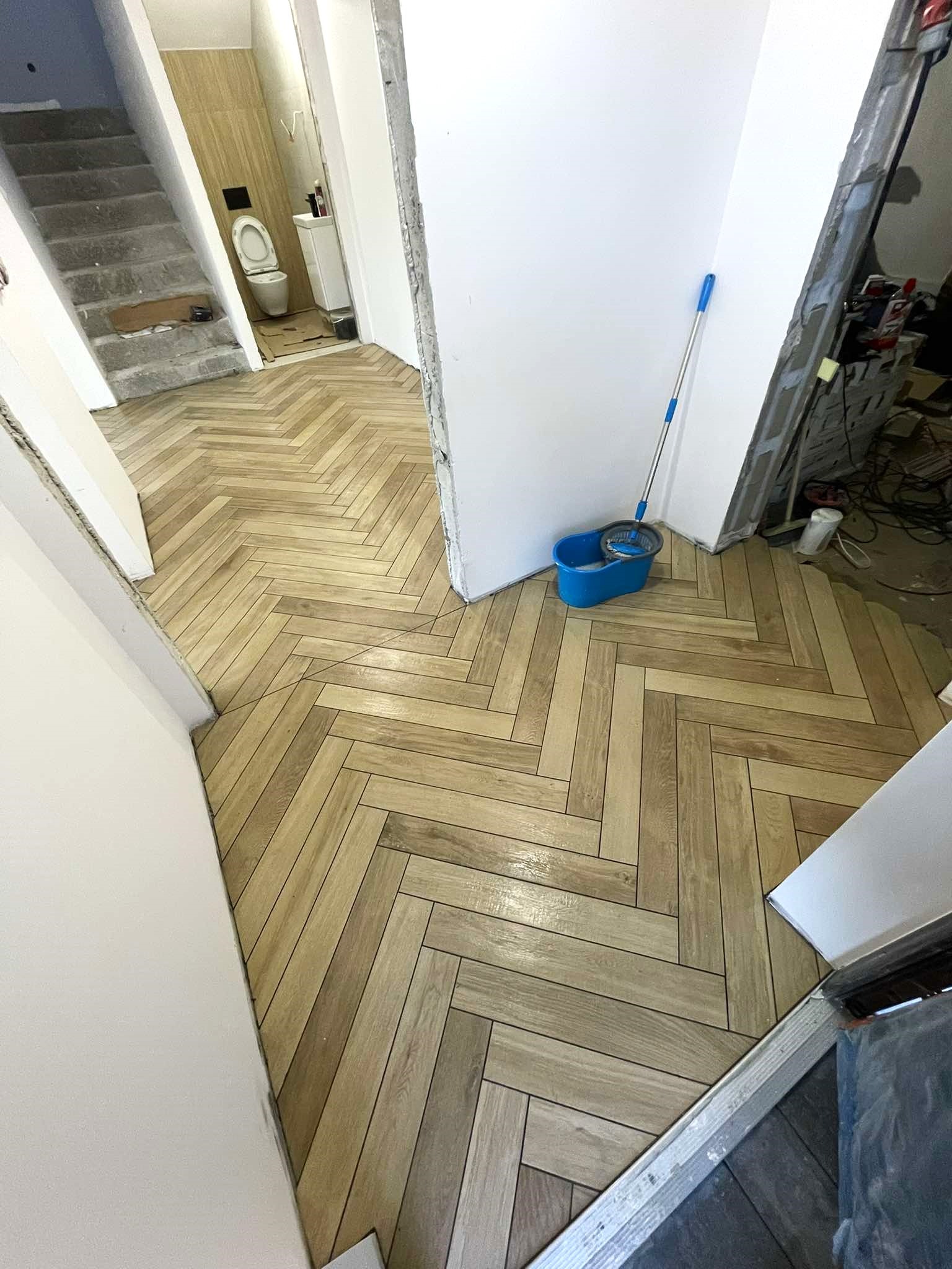 Ułożona w jodełkę, drewnopodobna glazura podłogowa w korytarzu i łazience, widoczny mop i wiadro, schody w tle.