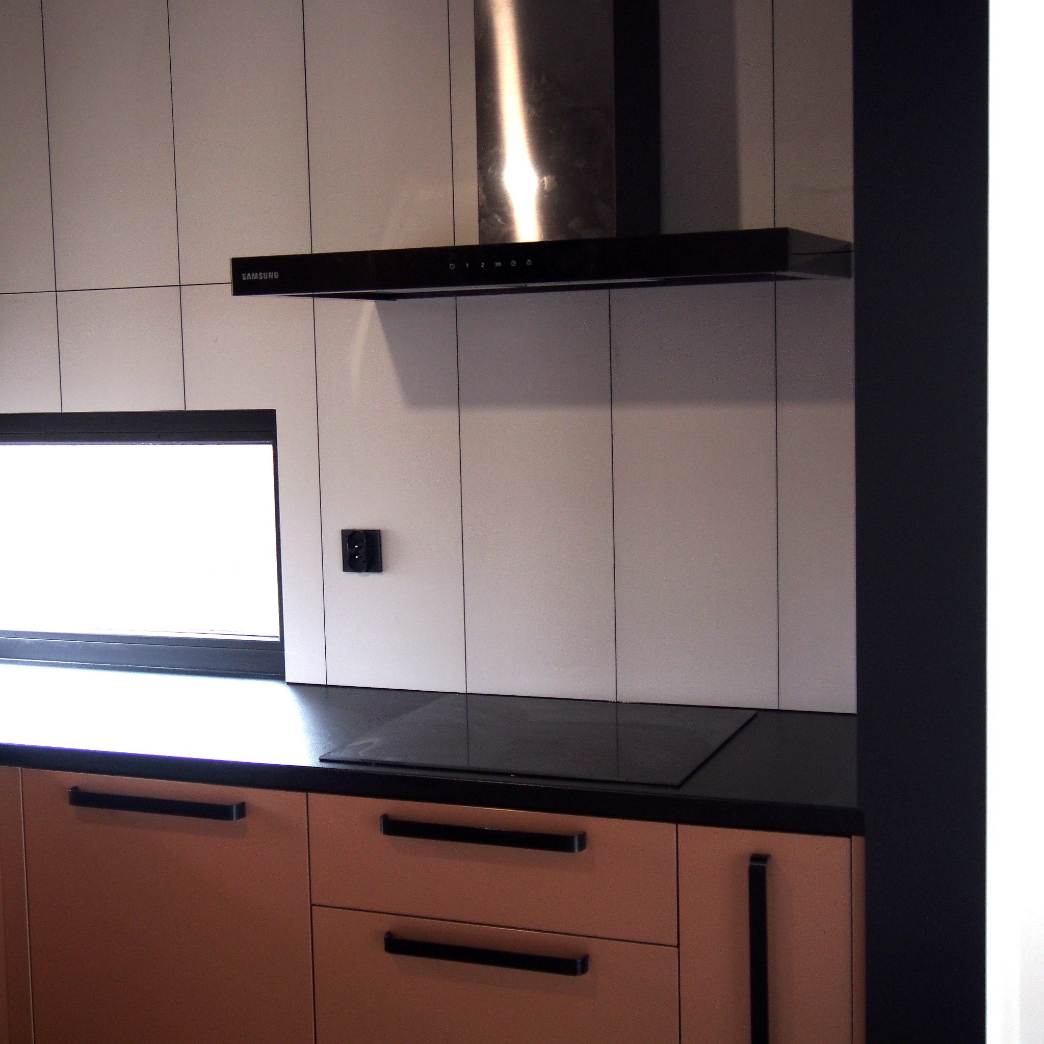 Nowoczesna kuchnia z beżowymi szafkami, czarnym blatem, płytą indukcyjną i okapem Samsung, wykończona białymi, kwadratowymi płytkami z czarnymi fugami i małym oknem.