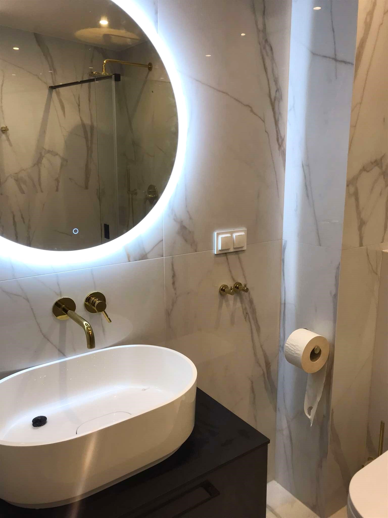 Nowoczesna łazienka z marmurowymi płytkami, owalną umywalką nablatową, złotą armaturą i okrągłym lustrem z podświetleniem LED.
