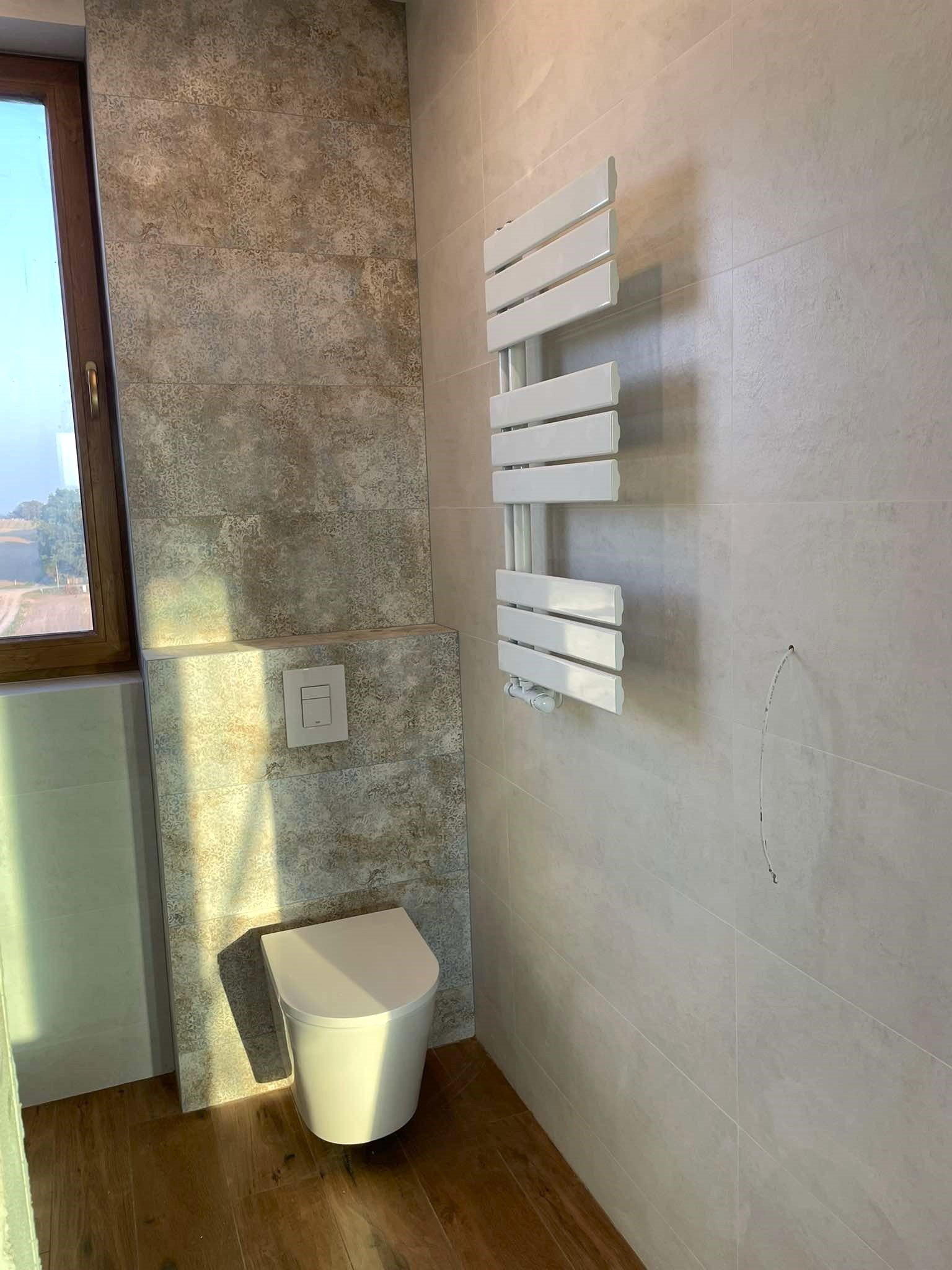 Nowoczesna łazienka z wiszącą toaletą, grzejnikiem drabinkowym i płytkami imitującymi kamień w odcieniach beżu i szarości, widok na krajobraz za oknem.