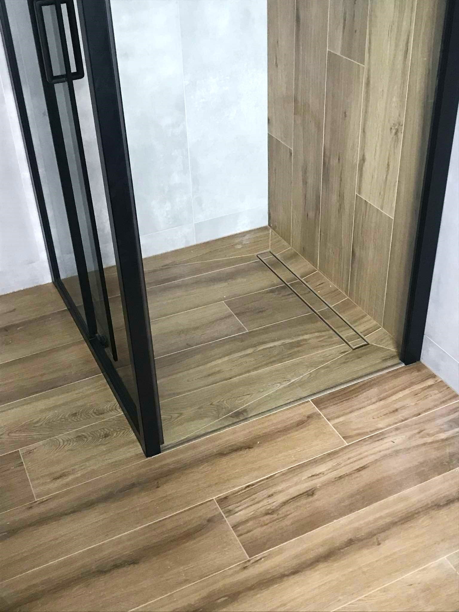 Nowoczesna kabina prysznicowa z czarnymi profilami i odpływem liniowym, wyłożona płytkami imitującymi drewno w odcieniach brązu i beżu, z widocznymi jasnymi płytkami na ścianie w tle.