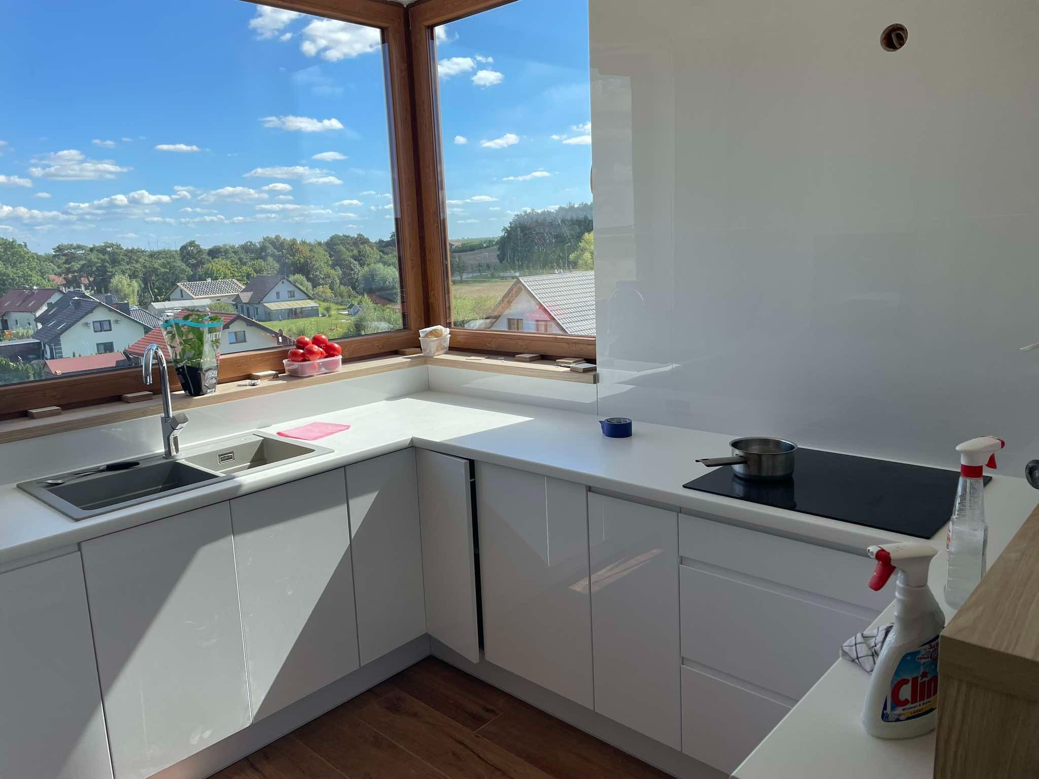 Jasna, nowoczesna kuchnia z widokiem na zielone otoczenie przez duże okno. Białe szafki, drewniana podłoga i czarna płyta grzewcza z garnkiem. Na blacie butelka płynu do czyszczenia.