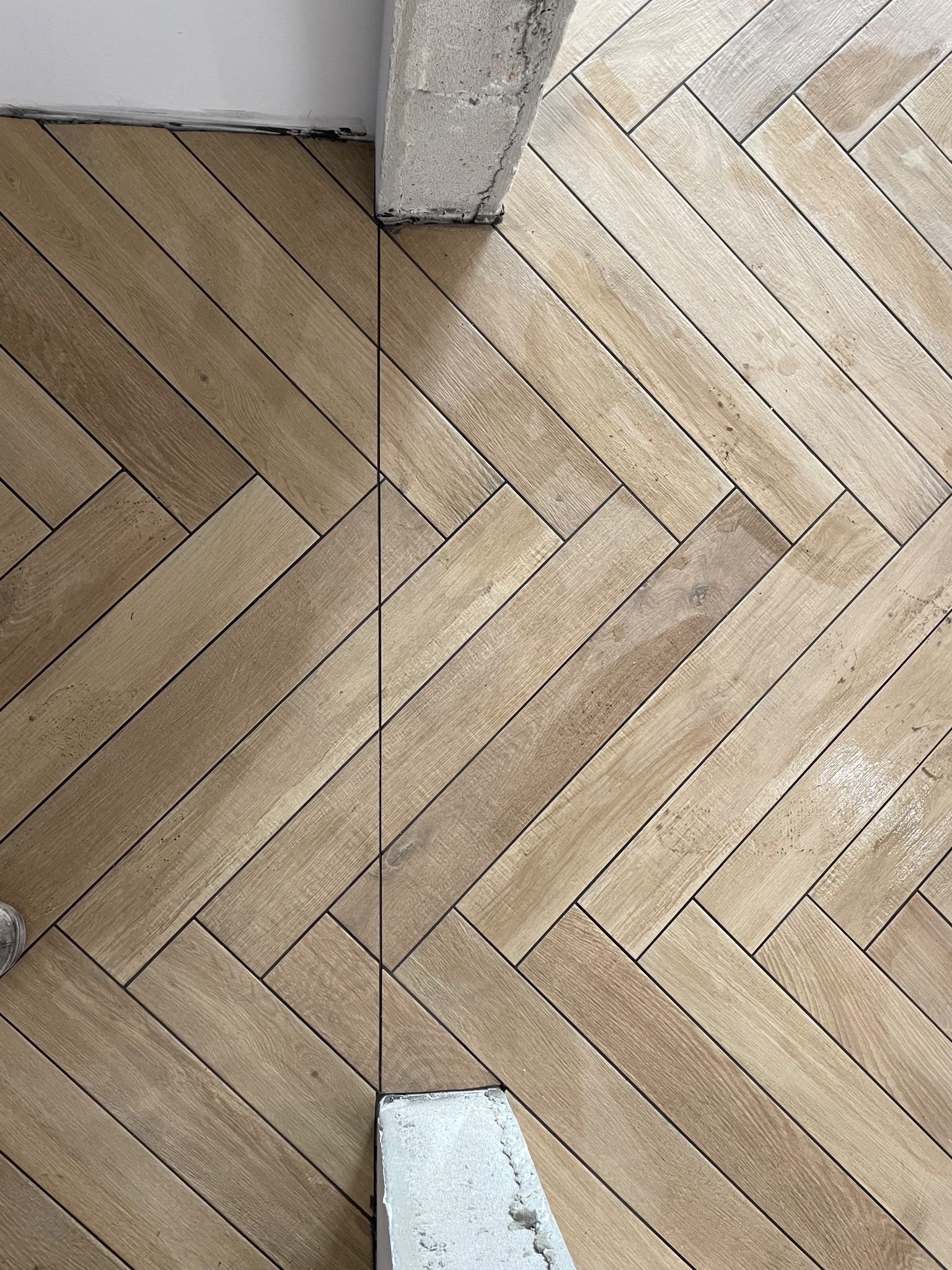 Ułożona w jodełkę jasnobrązowa podłoga z płytek imitujących drewno, z widocznym fragmentem ściany i słupa w trakcie prac wykończeniowych.