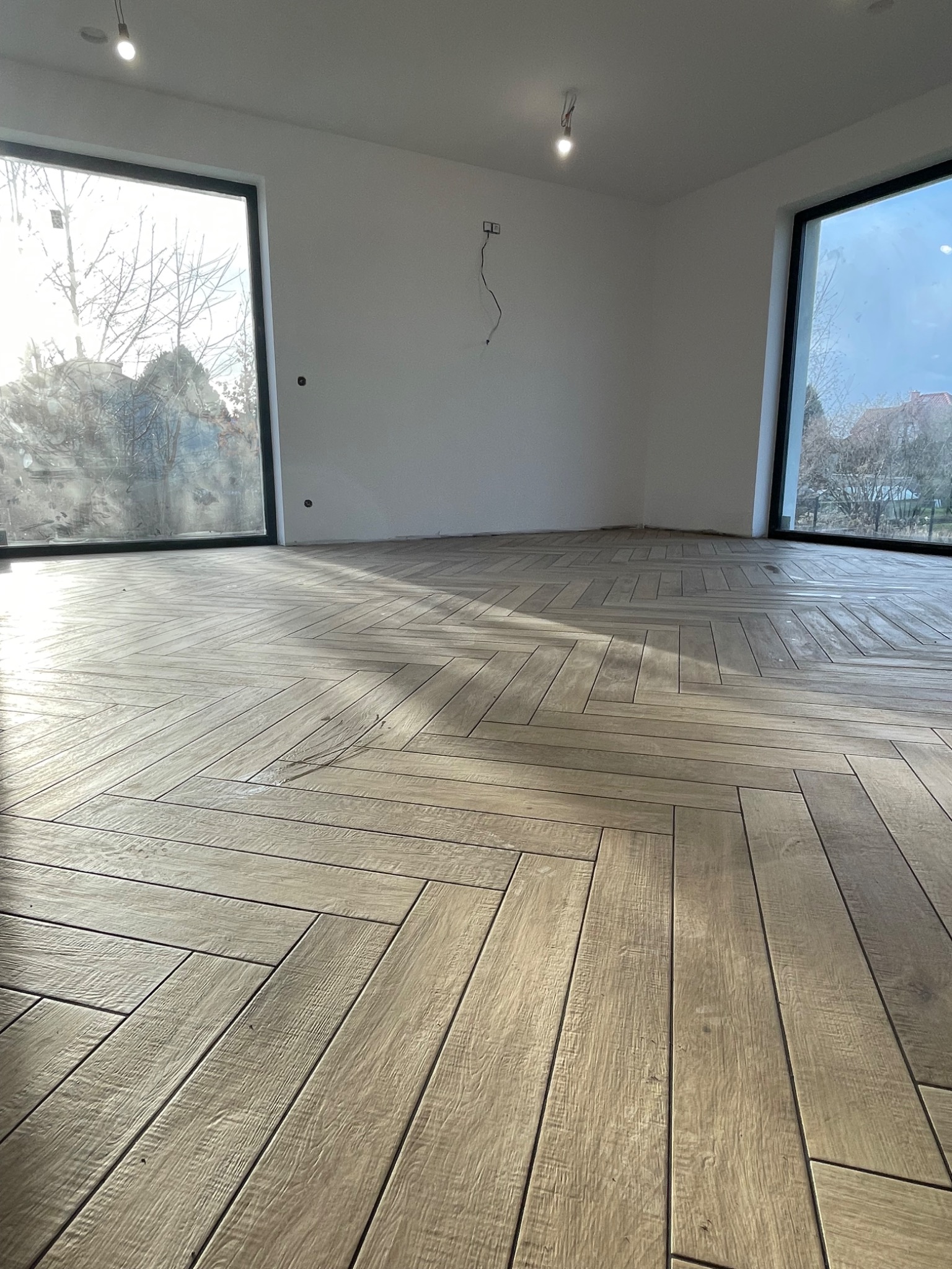 Ułożona w jodełkę podłoga z płytek imitujących drewno w jasnym, pustym pokoju z dużymi oknami i minimalistycznym oświetleniem.