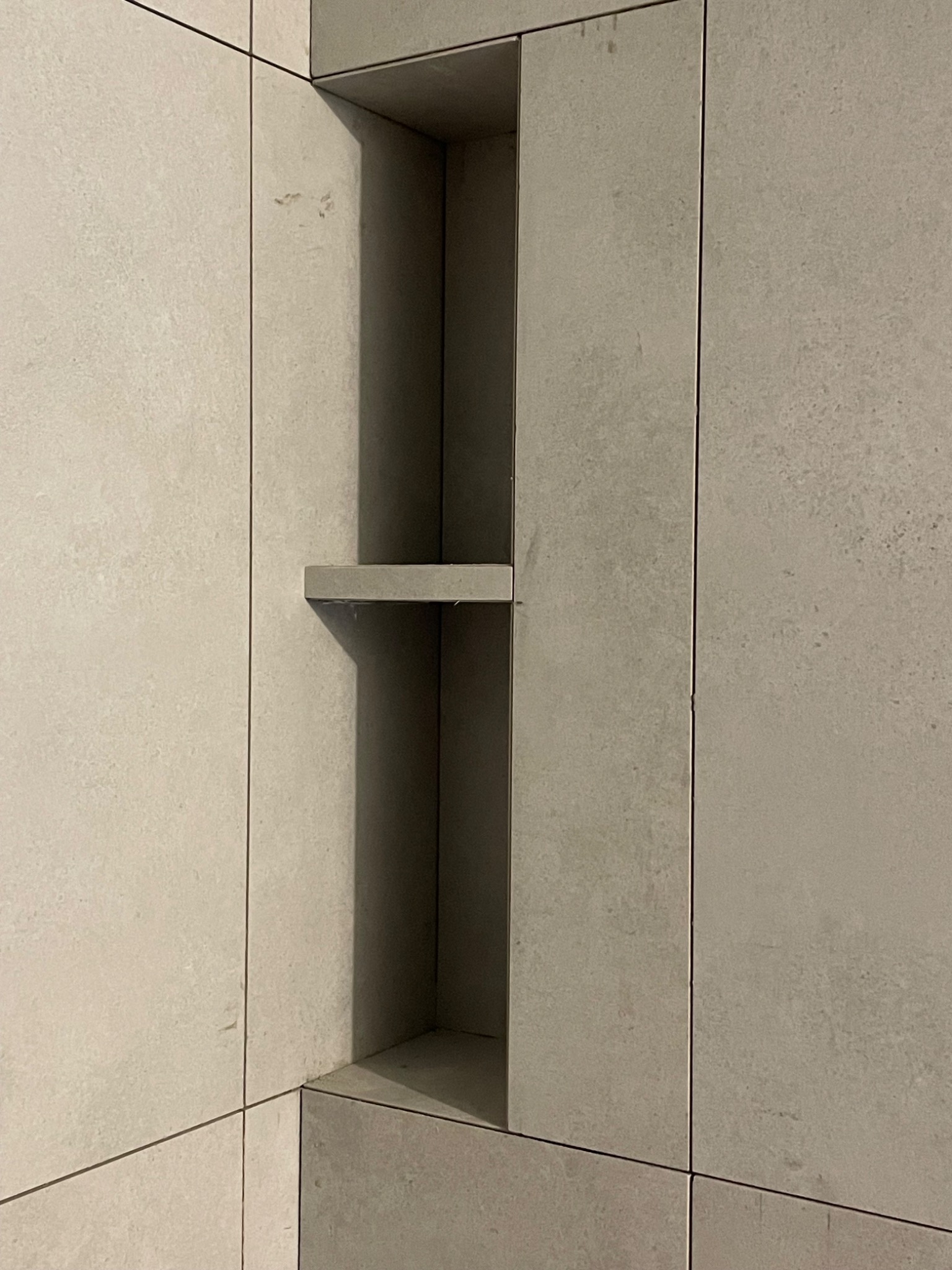 Wnęka z półką wyłożona płytkami w odcieniach szarości, z widocznymi fugami, stanowiąca element aranżacji ściany w łazience.