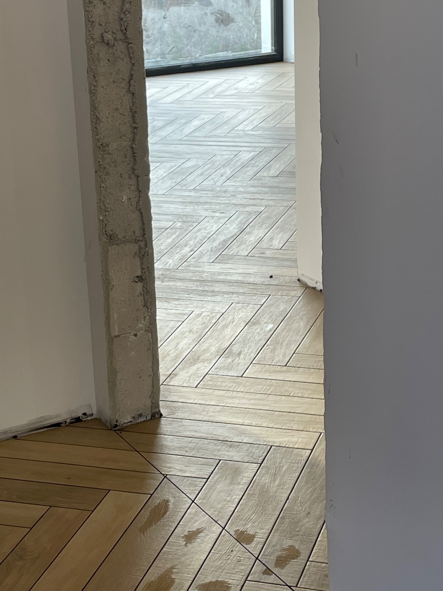 Ułożona w jodełkę, drewnopodobna podłoga z płytek w Malborku, widoczna przez surowy, betonowy filar w trakcie wykańczania wnętrza.