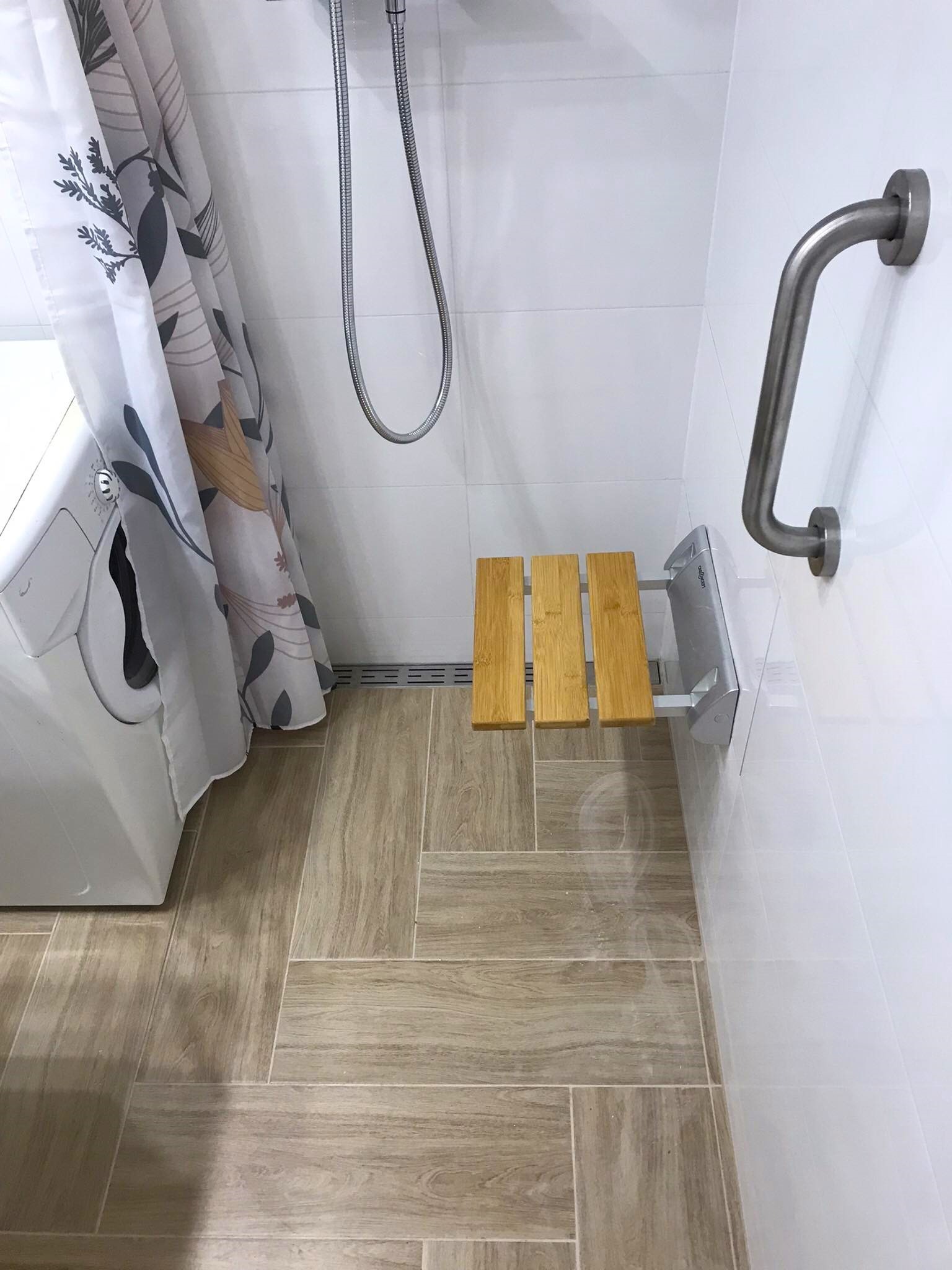 Biała łazienka z drewnianą składaną ławeczką prysznicową i uchwytem dla niepełnosprawnych, płytki podłogowe imitujące drewno, zasłona w kwiaty, pralka z boku.