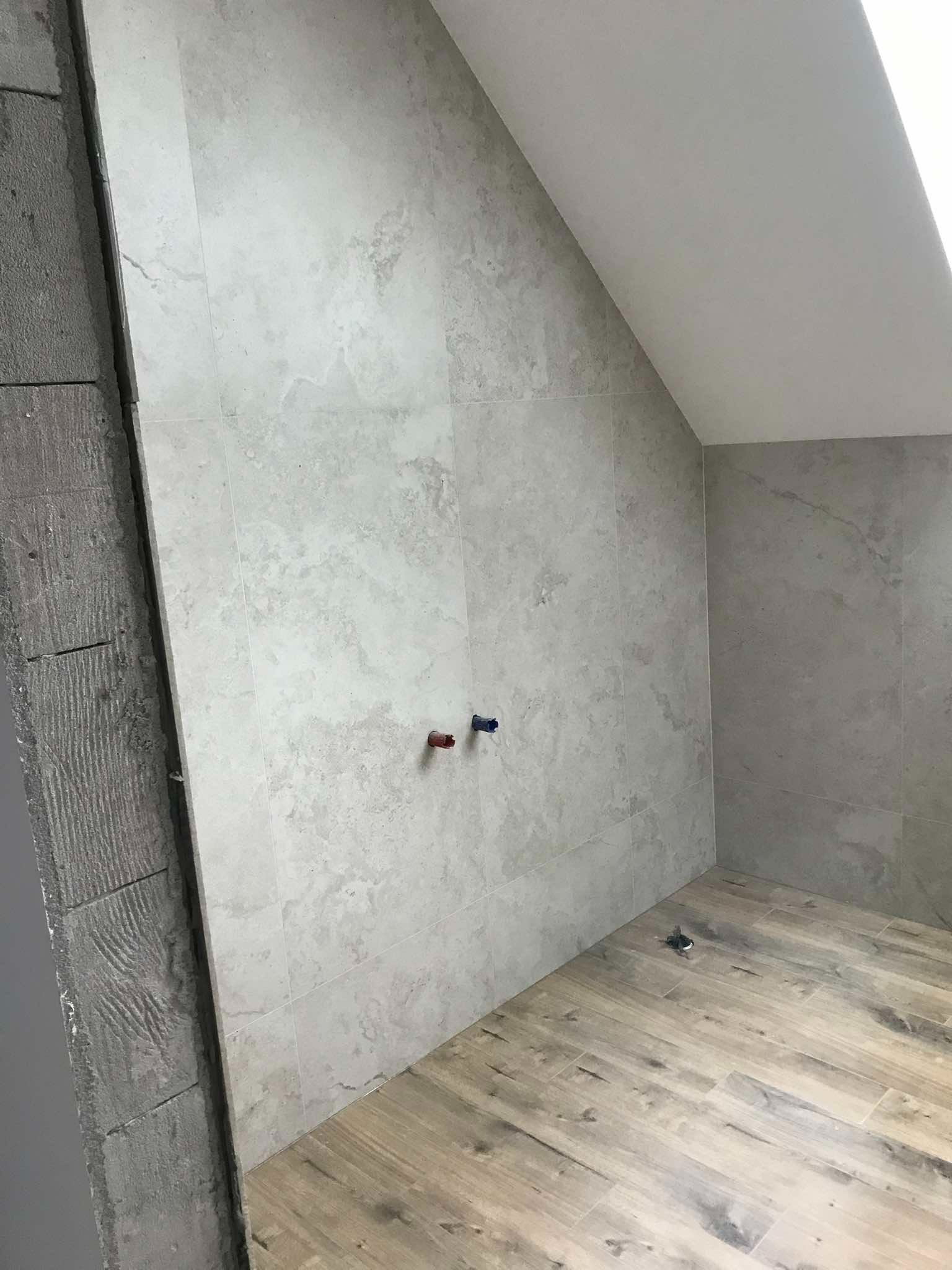 Łazienka w trakcie remontu: ściany wyłożone jasnoszarymi płytkami imitującymi kamień, podłoga z paneli drewnopodobnych, widoczne przyłącza wody i surowa ściana w trakcie przygotowania pod wykończenie.