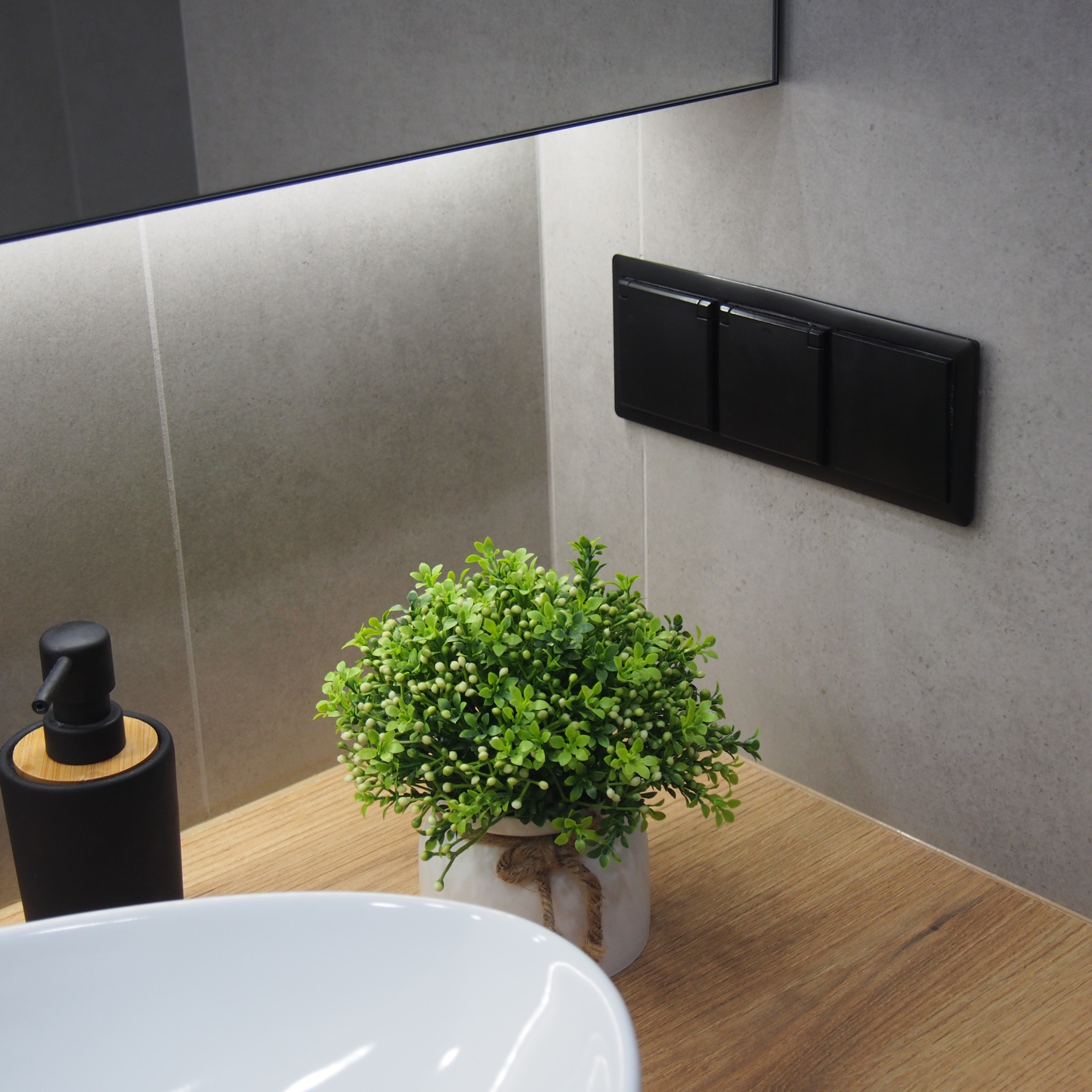 Nowoczesna łazienka z szarymi płytkami, drewnianym blatem, białą umywalką, czarnym dozownikiem mydła i potrójnym czarnym włącznikiem światła, oświetlona listwą LED pod lustrem, z dekoracyjną...