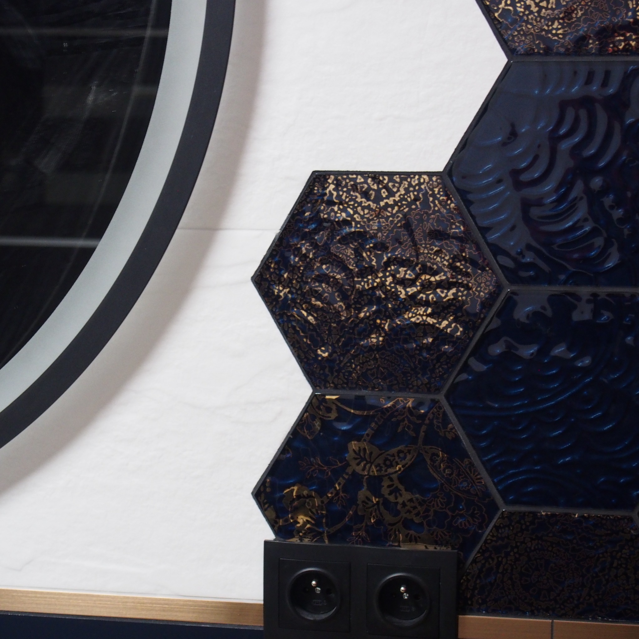 Fragment ściany łazienki z heksagonalnymi, granatowymi płytkami z wzorami w odcieniach złota, obok okrągłe lustro z czarną ramą i podwójne gniazdko elektryczne w czarnej obudowie.