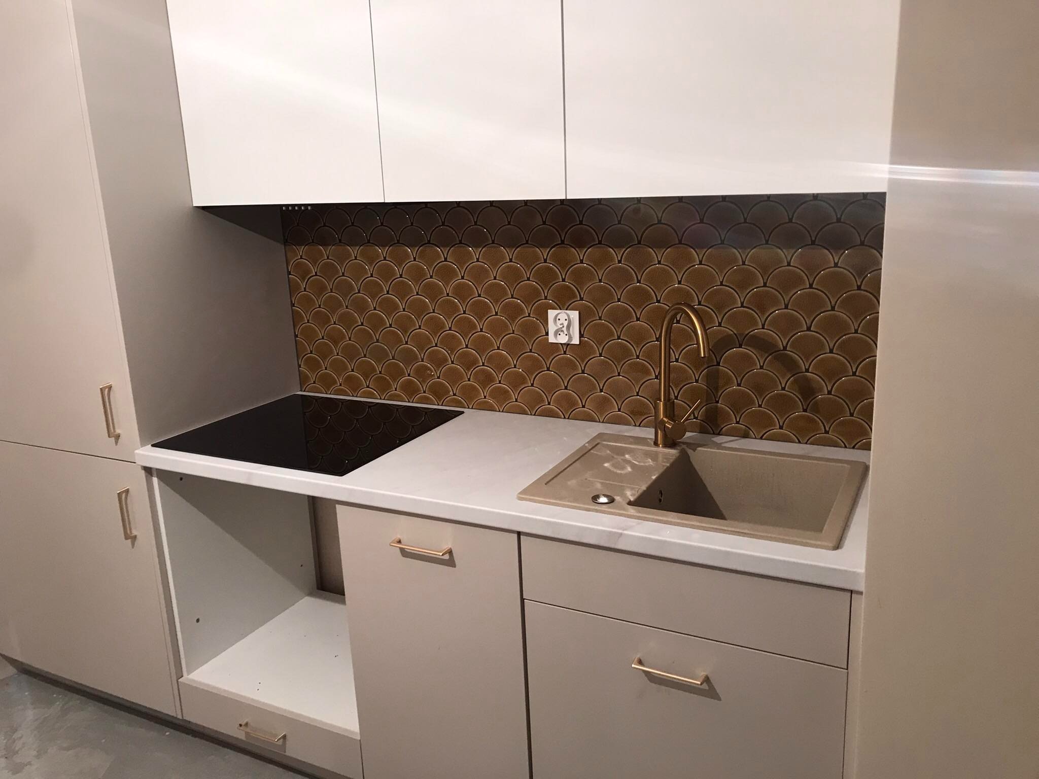 Nowoczesna kuchnia z brązową mozaiką w kształcie rybiej łuski nad blatem, złota bateria przy beżowym zlewozmywaku i białe szafki z podłużnymi złotymi uchwytami.