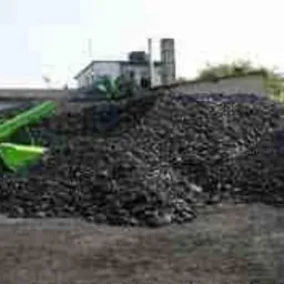Góra czarnego węgla na placu, z widocznym w tle zielonym wózkiem widłowym i budynkiem.