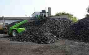 Góra czarnego węgla na placu, z widocznym w tle zielonym wózkiem widłowym i budynkiem.