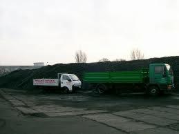 Dwa samochody ciężarowe z otwartą skrzynią ładunkową stojące na placu przed hałdą węgla. Jeden pojazd biały, drugi zielony. Niebo pochmurne.