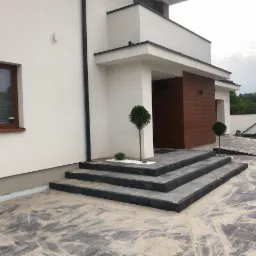 Nowoczesny dom jednorodzinny z elewacją w kolorze złamanej bieli, wejściem wykończonym drewnem, schodami z szarej kostki z czarnymi obrzeżami i podjazdem z tej samej kostki, częściowo zasypanym...