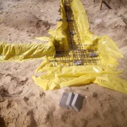 Przygotowanie wykopu pod fundament z żółtą folią ochronną i zbrojeniem z prętów stalowych; w tle widoczne drewniane słupki wyznaczające granice.