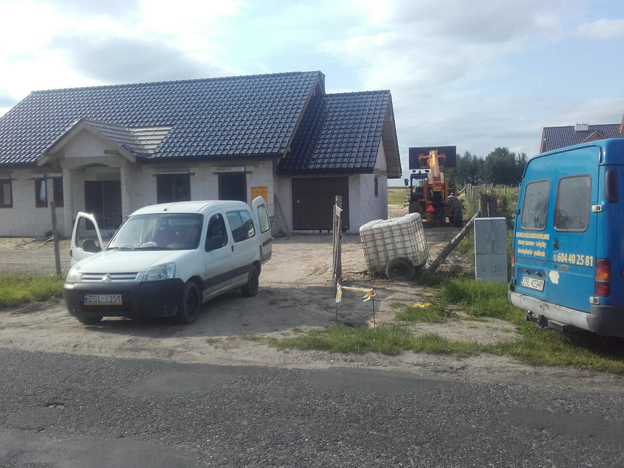 Niedokończony dom jednorodzinny z ciemną dachówką, biały samochód dostawczy z otwartymi drzwiami i niebieski samochód dostawczy z reklamą firmy budowlanej w tle, na placu budowy. Widoczny sprzęt...