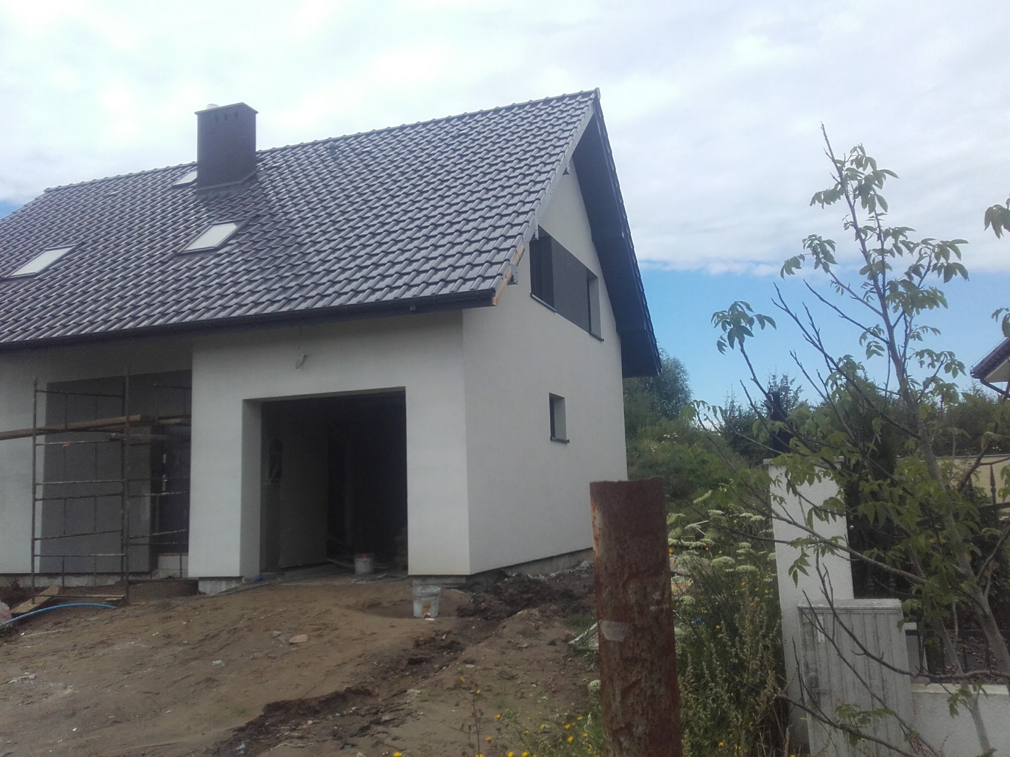 Surowy, biały dom jednorodzinny z ciemną dachówką i lukarnami, widoczny w trakcie budowy, z rusztowaniem z boku i ziemią wokół fundamentów.