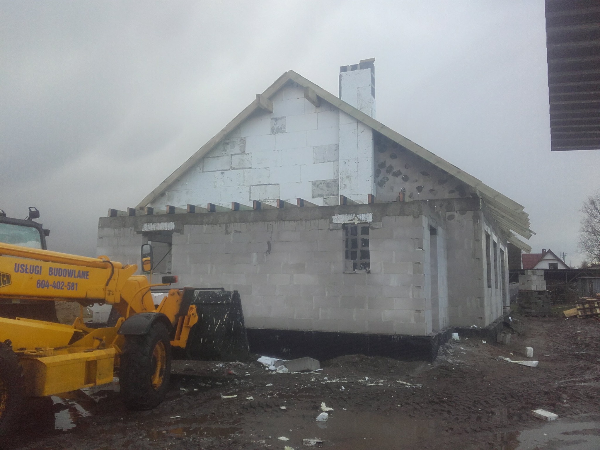 Surowy dom w trakcie budowy, ściany z szarego betonu komórkowego, ocieplony szczyt dachu, na pierwszym planie żółta ładowarka teleskopowa z widocznym numerem telefonu.