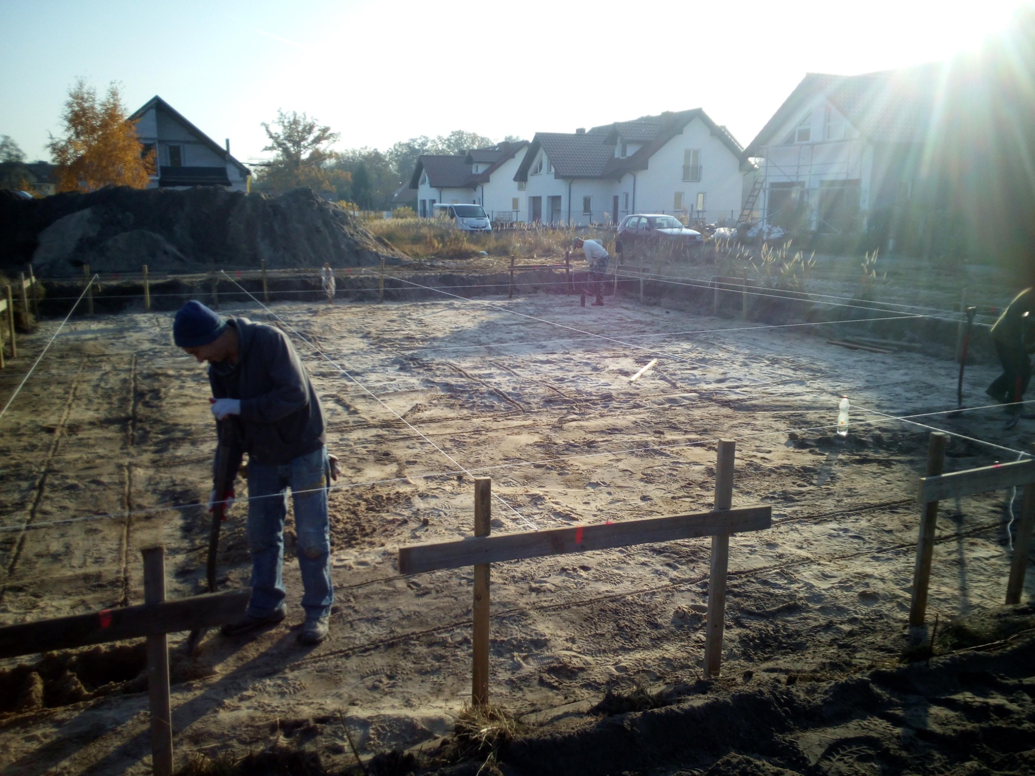 Pracownik w niebieskiej czapce wbija kołek w ziemię na placu budowy, wyznaczając granice fundamentów pod budynek mieszkalny; w tle widoczne są kolejne domy w różnym stadium budowy.