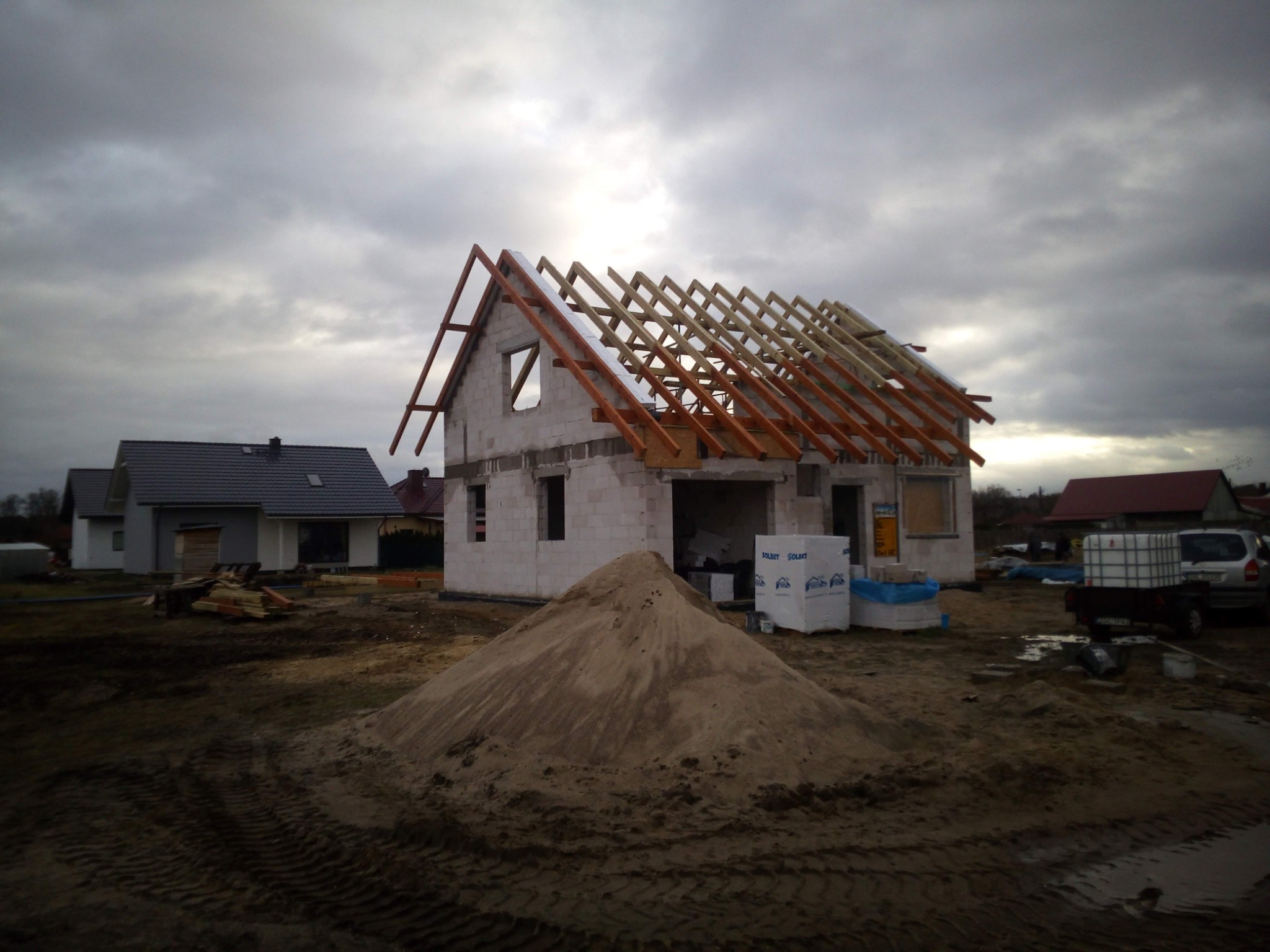 Surowy stan domu z pustaków, widoczna konstrukcja dachu z drewna, sterta piasku na pierwszym planie, budowa w toku, pochmurne niebo w tle.