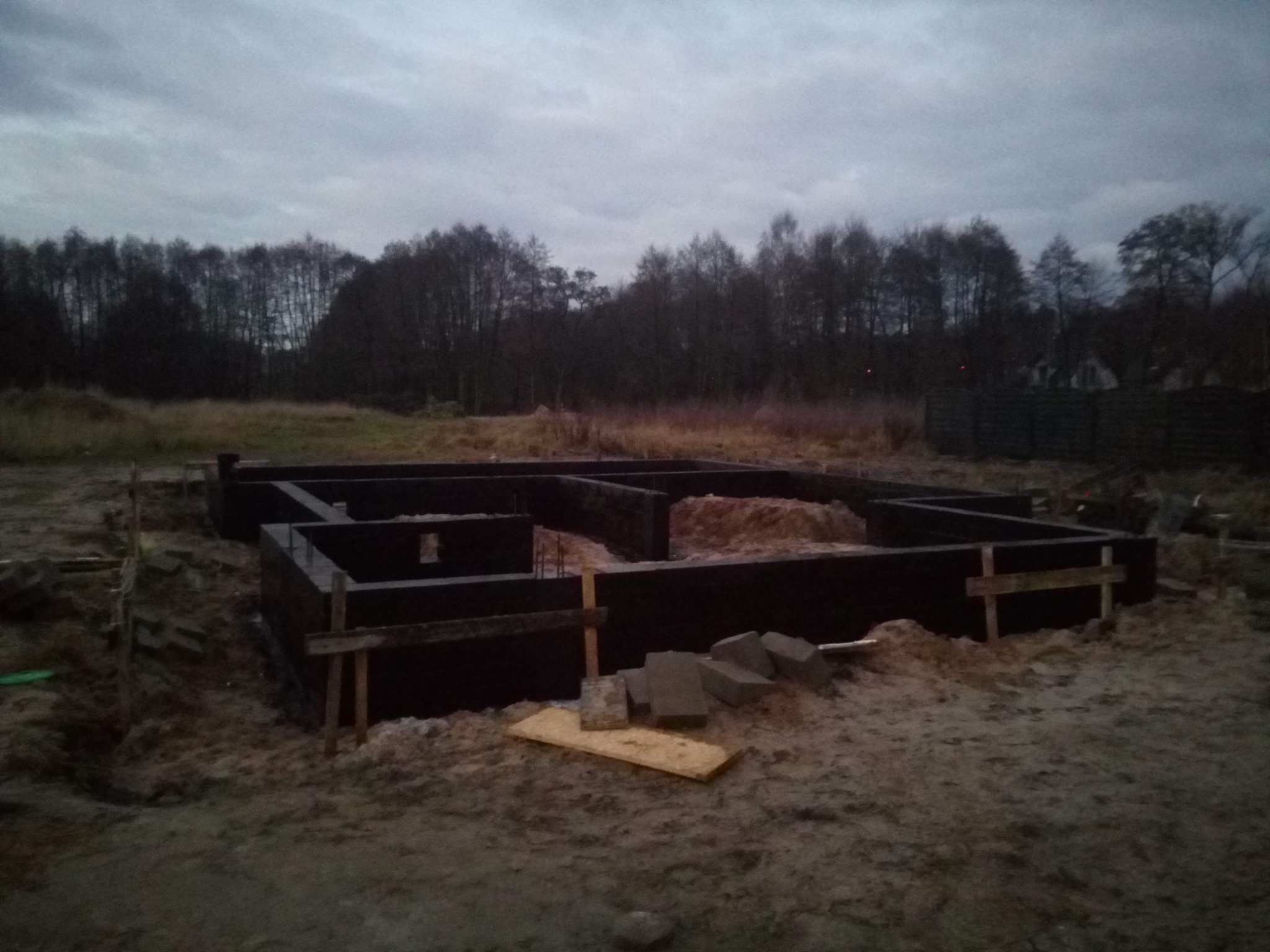 Wylane fundamenty pod budynek z widocznymi deskami szalunkowymi i częściowo zasypanym piaskiem wewnątrz, na tle pochmurnego nieba i drzew.