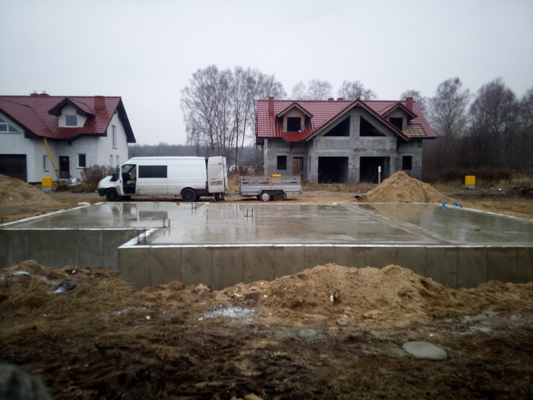 Wylana i mokra płyta fundamentowa pod budowę domu murowanego, z widocznym szalunkiem i wystającymi prętami zbrojeniowymi, na tle surowego stanu budynku w trakcie budowy i zaparkowanego białego busa...