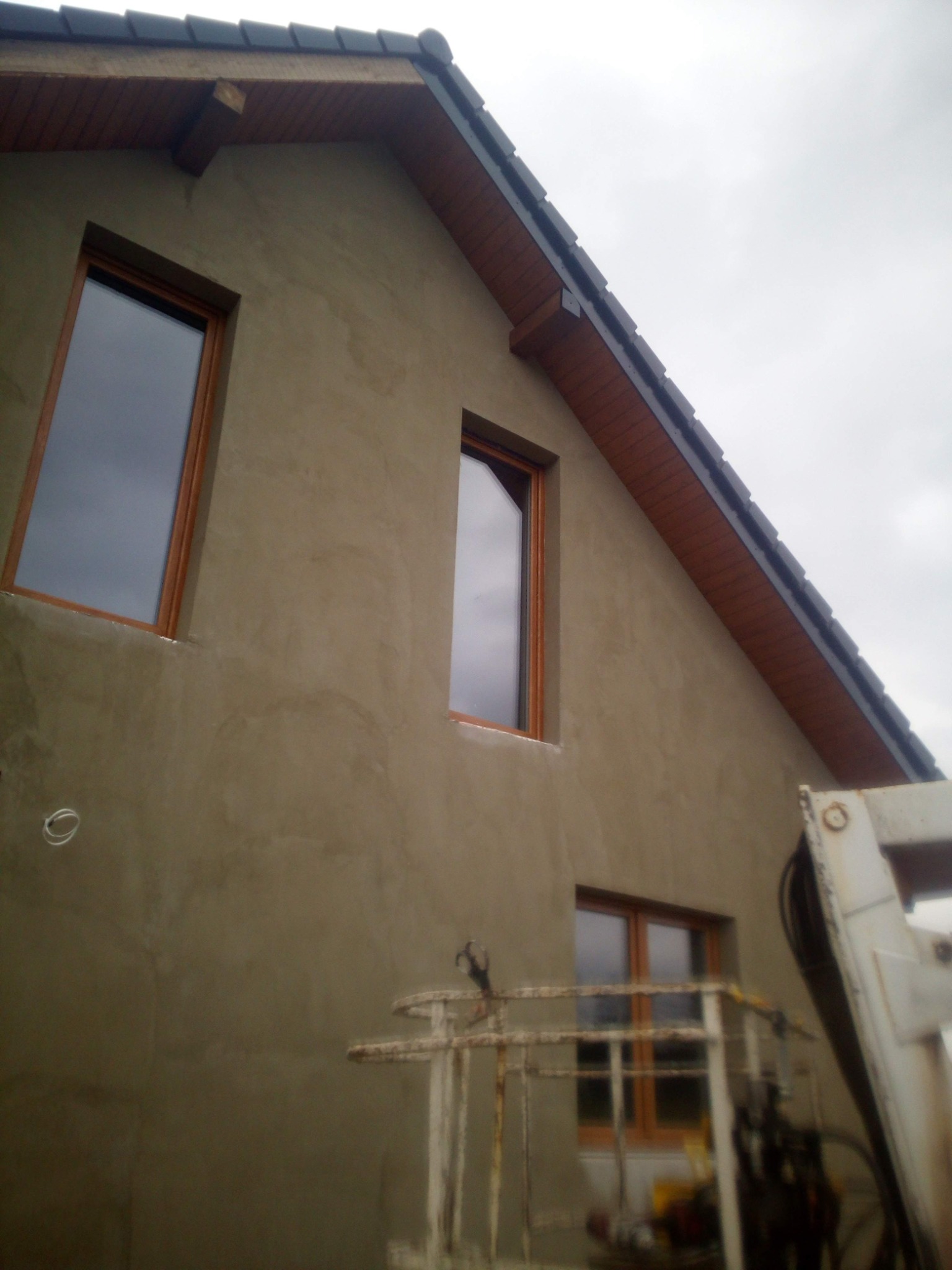 Ściana domu w trakcie tynkowania, widoczne okna z drewnianą stolarką i fragment podnośnika koszowego.