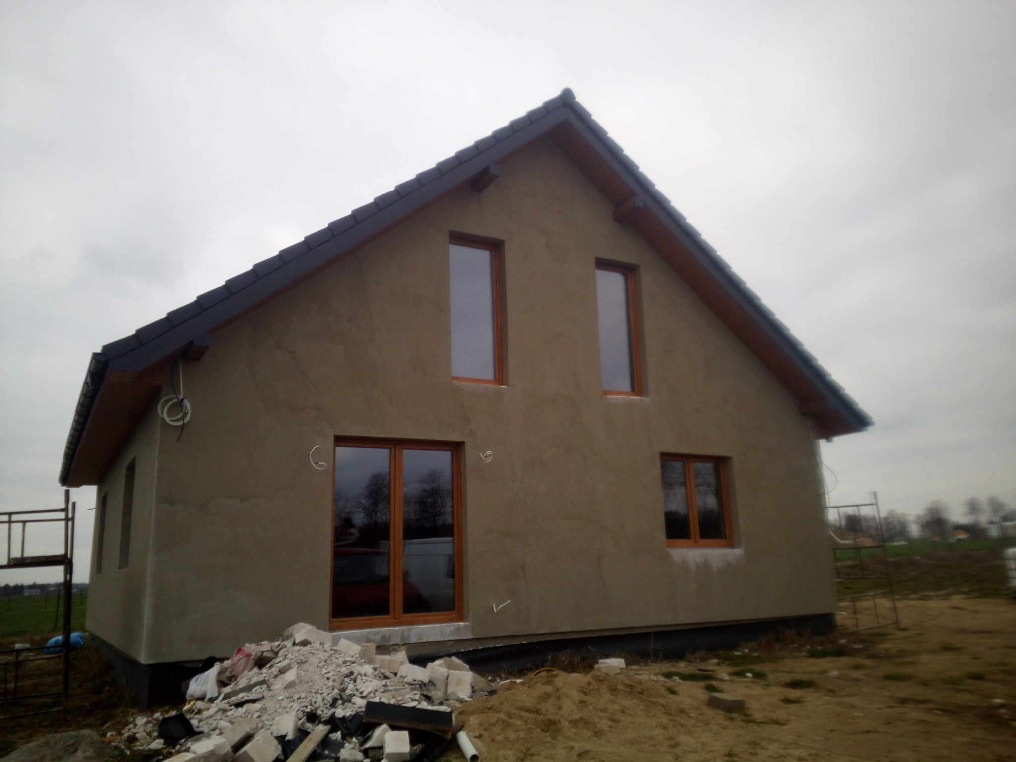 Surowy dom murowany z brązowymi oknami, otynkowany szarą zaprawą, z czarną dachówką, widok z zewnątrz na plac budowy z gruzem i piaskiem.