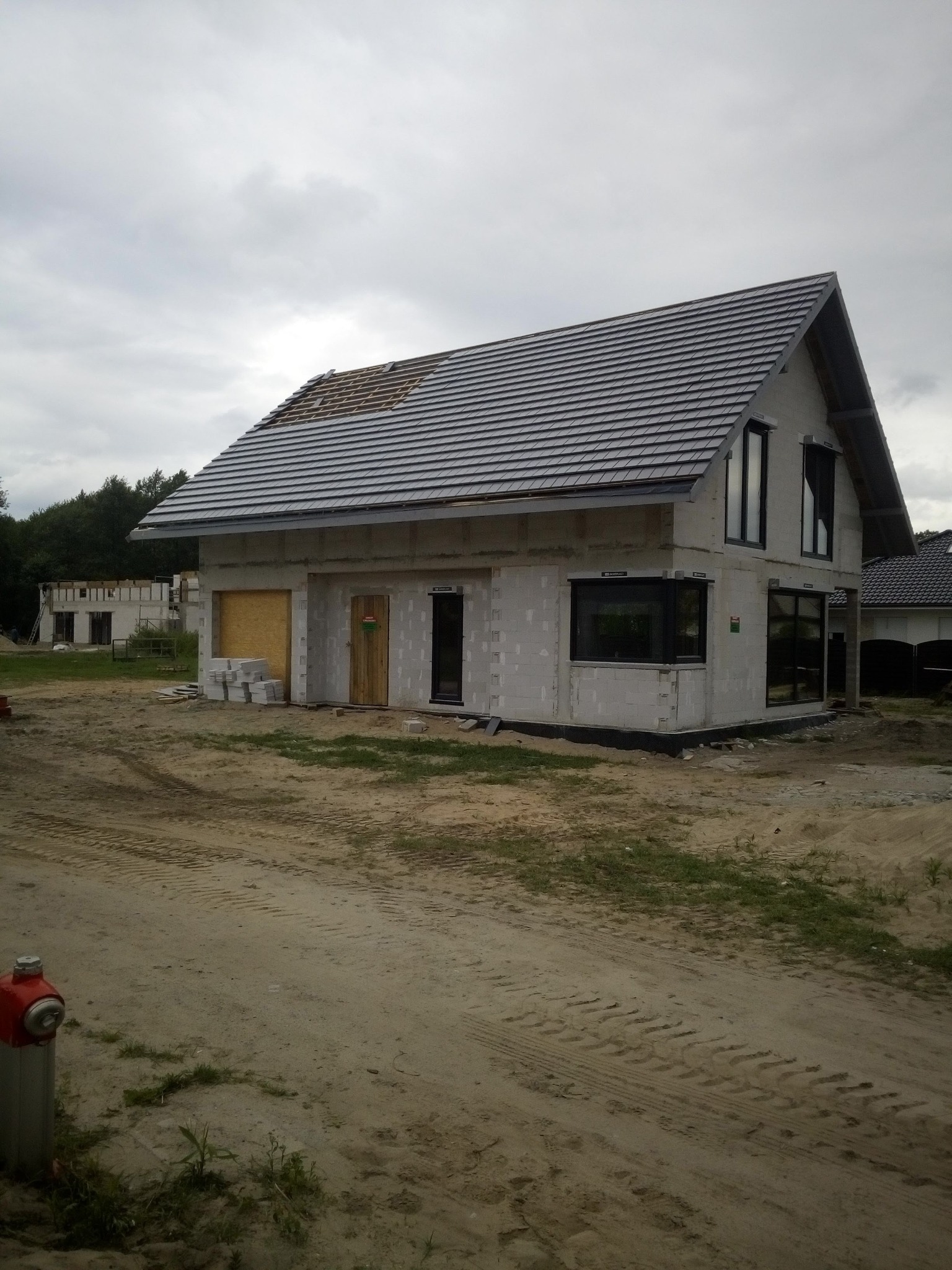 Surowy dom murowany w trakcie budowy z częściowo ułożonym dachem z szarej dachówki, widoczne pustaki, okna z czarną ramą i prowizoryczne drzwi z płyty, na pierwszym planie piaszczysta droga.
