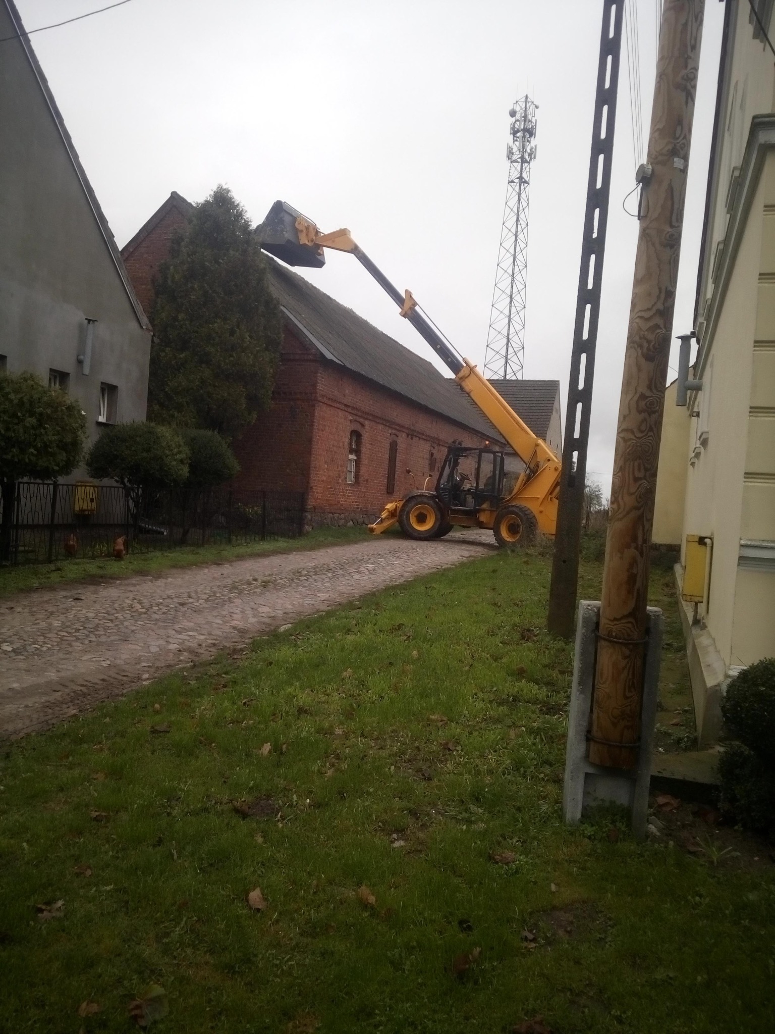 Żółta ładowarka teleskopowa z podniesionym ramieniem i łyżką na tle ceglanego budynku z szarym dachem i wieżą telekomunikacyjną w pochmurny dzień.