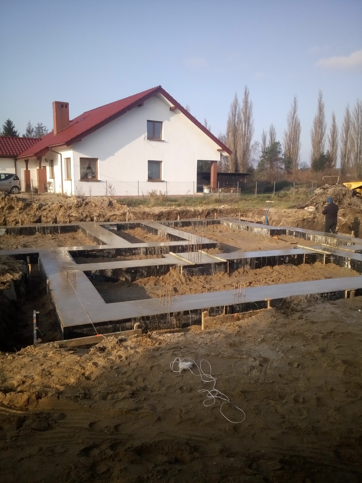 Wylane fundamenty pod budowę domu jednorodzinnego z widocznym zbrojeniem i częściowo wykopanym terenem, na drugim planie biały dom z czerwoną dachówką i drzewa.