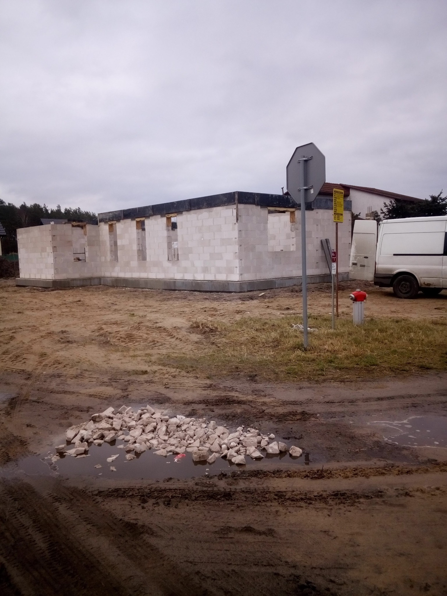 Surowy stan budowy domu z białych bloczków, widoczne otwory okienne i drzwiowe, teren budowy błotnisty, z kupką gruzu w kałuży na pierwszym planie, zaparkowane busy w tle.