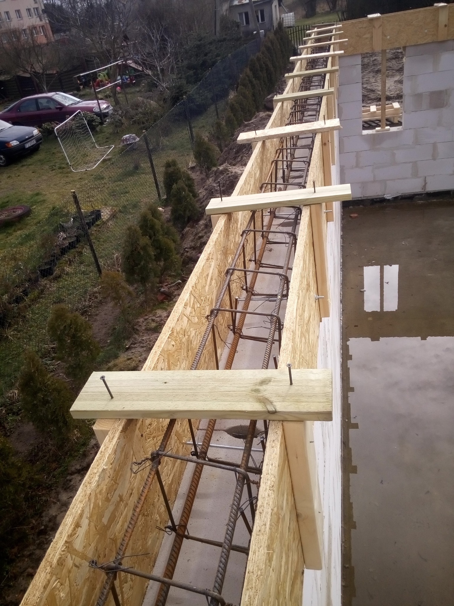 Szczegółowe ujęcie szalunku z drewnianych płyt OSB i belek, zbrojenia stalowego oraz fragmentu ściany z bloczków betonowych na budowie domu jednorodzinnego, z widocznym odbiciem w kałuży...