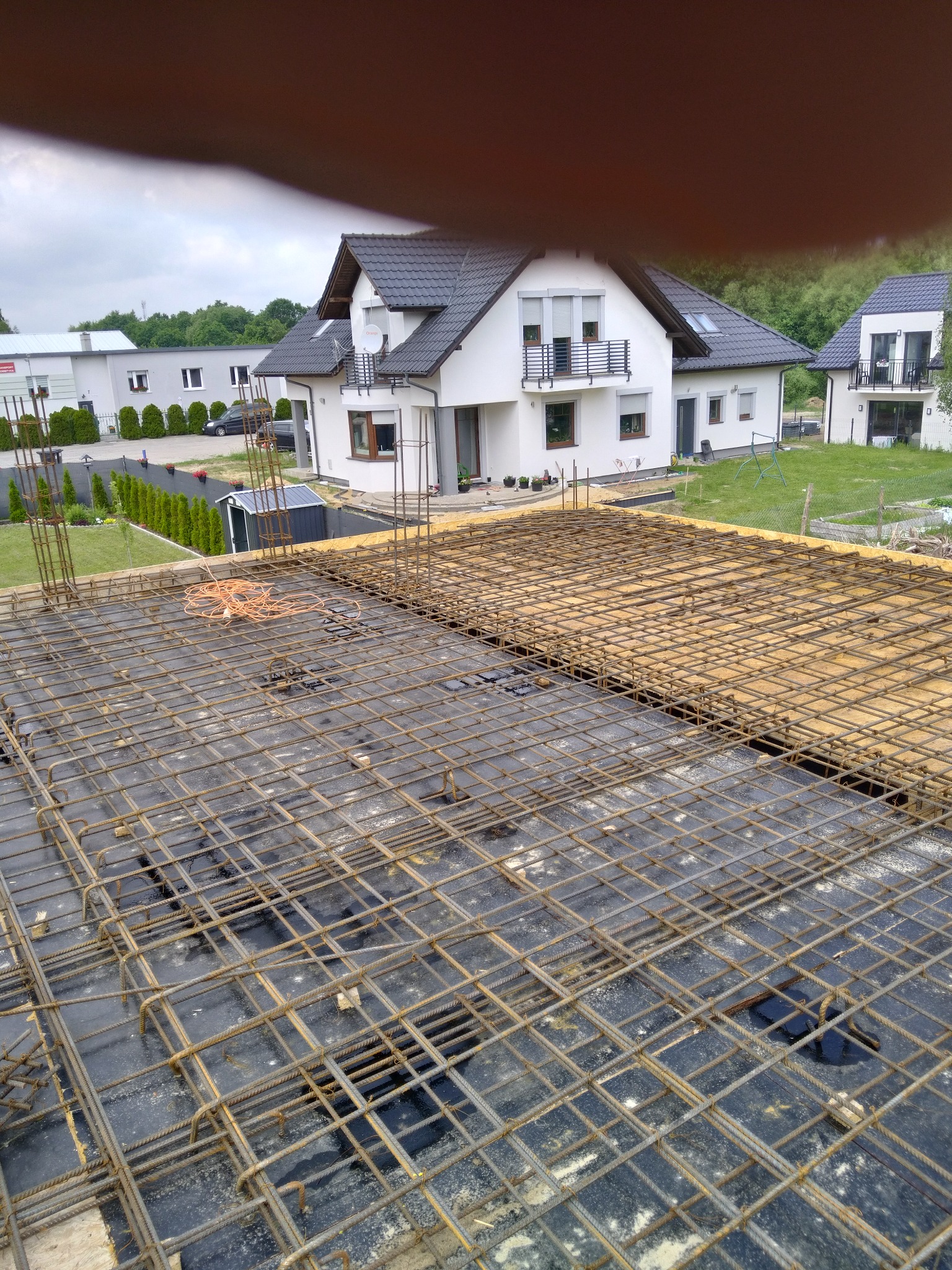 Zbrojenie płyty fundamentowej z widocznymi prętami stalowymi na czarnej folii, w tle dwa białe domy z ciemnymi dachami i zielony trawnik.