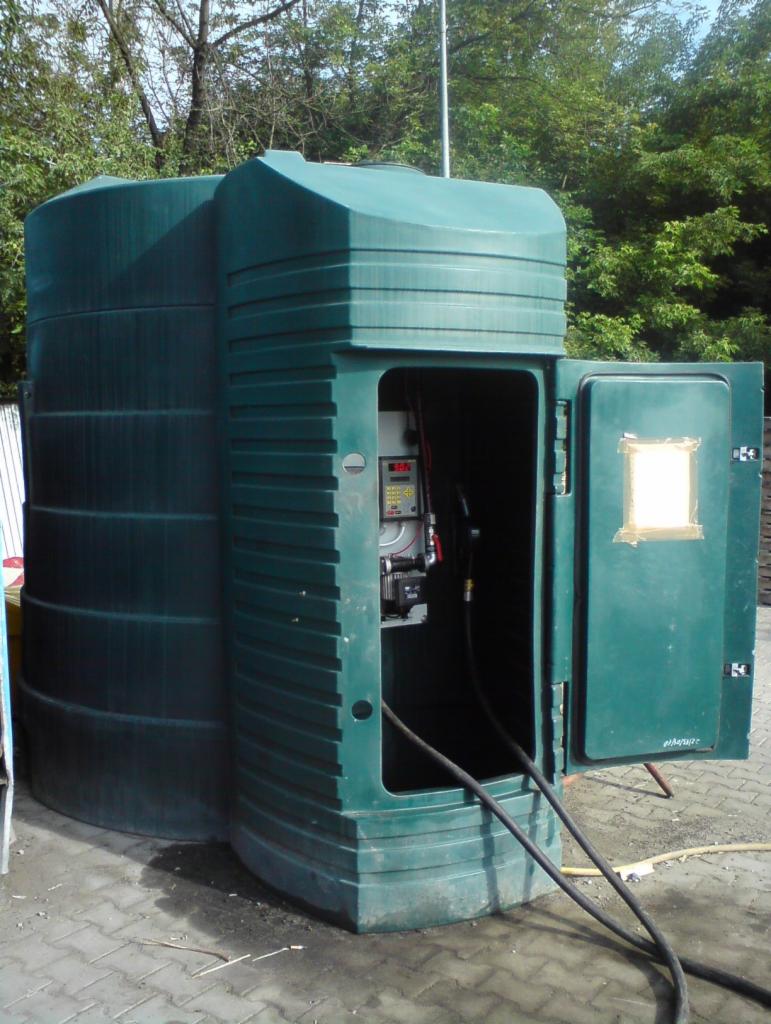 Zielony, dwukomorowy zbiornik na olej napędowy z otwartymi drzwiczkami, ukazującymi wewnętrzny mechanizm dystrybucji paliwa i dwa czarne węże leżące na kostce brukowej.