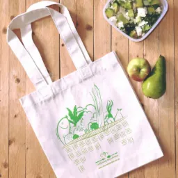 Biała torba bawełniana z zielonym nadrukiem warzyw, jabłko, gruszka i pojemnik z sałatką na drewnianym stole.