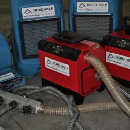 Profesjonalne urządzenia do osuszania budynków: niebieskie dmuchawy i czerwone kondensacyjne osuszacze z podłączonymi wężami, widoczne logo firmy Nord-Help.
