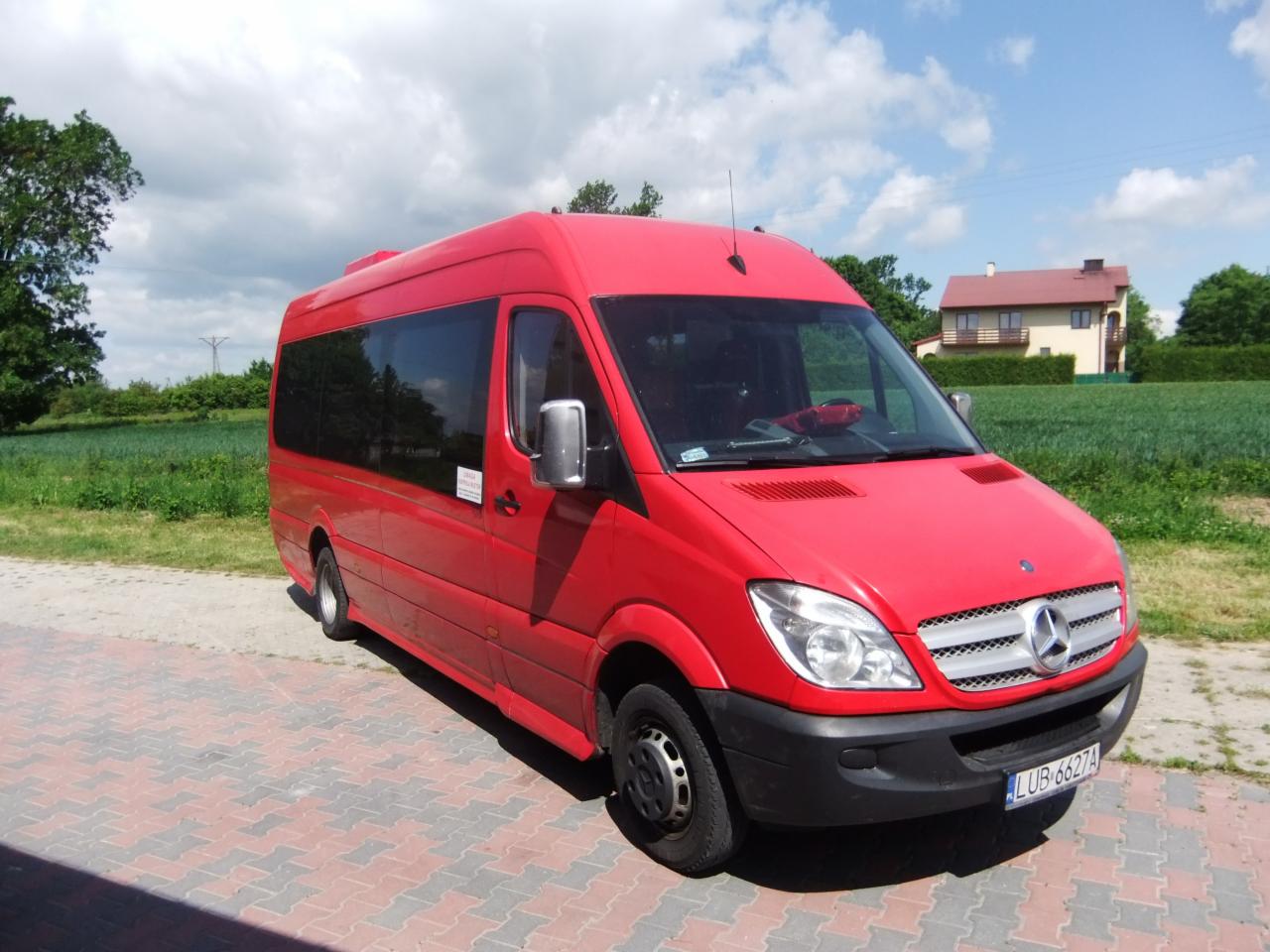 Czerwony bus Mercedes-Benz Sprinter z przyciemnianymi szybami, zaparkowany na kostce brukowej, z widocznym numerem rejestracyjnym LUB 6627A i domem w tle.