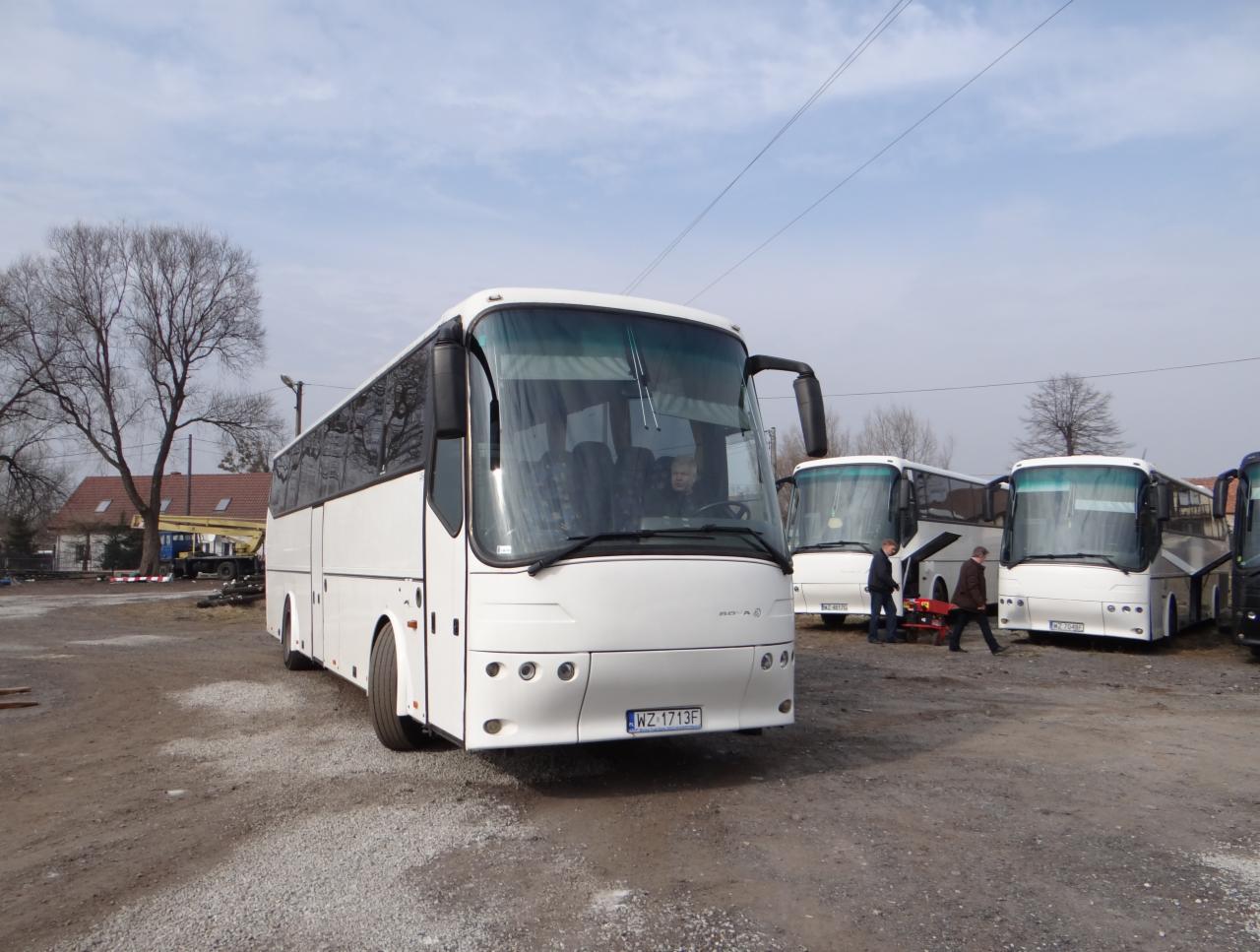 Flota białych autokarów na żwirowym placu, widoczny fragment budynku w tle, tablica rejestracyjna WZ 1713F na pierwszym planie.