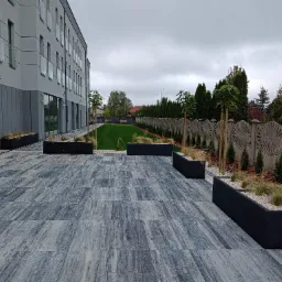 Nowoczesna aranżacja przestrzeni przed budynkiem z szarej kostki brukowej, czarnych donic z ozdobnymi trawami i zielonym trawnikiem w tle.