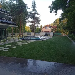 Zadbany trawnik z systemem nawadniania, płytami chodnikowymi prowadzącymi do zadaszonego basenu, widok na dom z ogrodem.